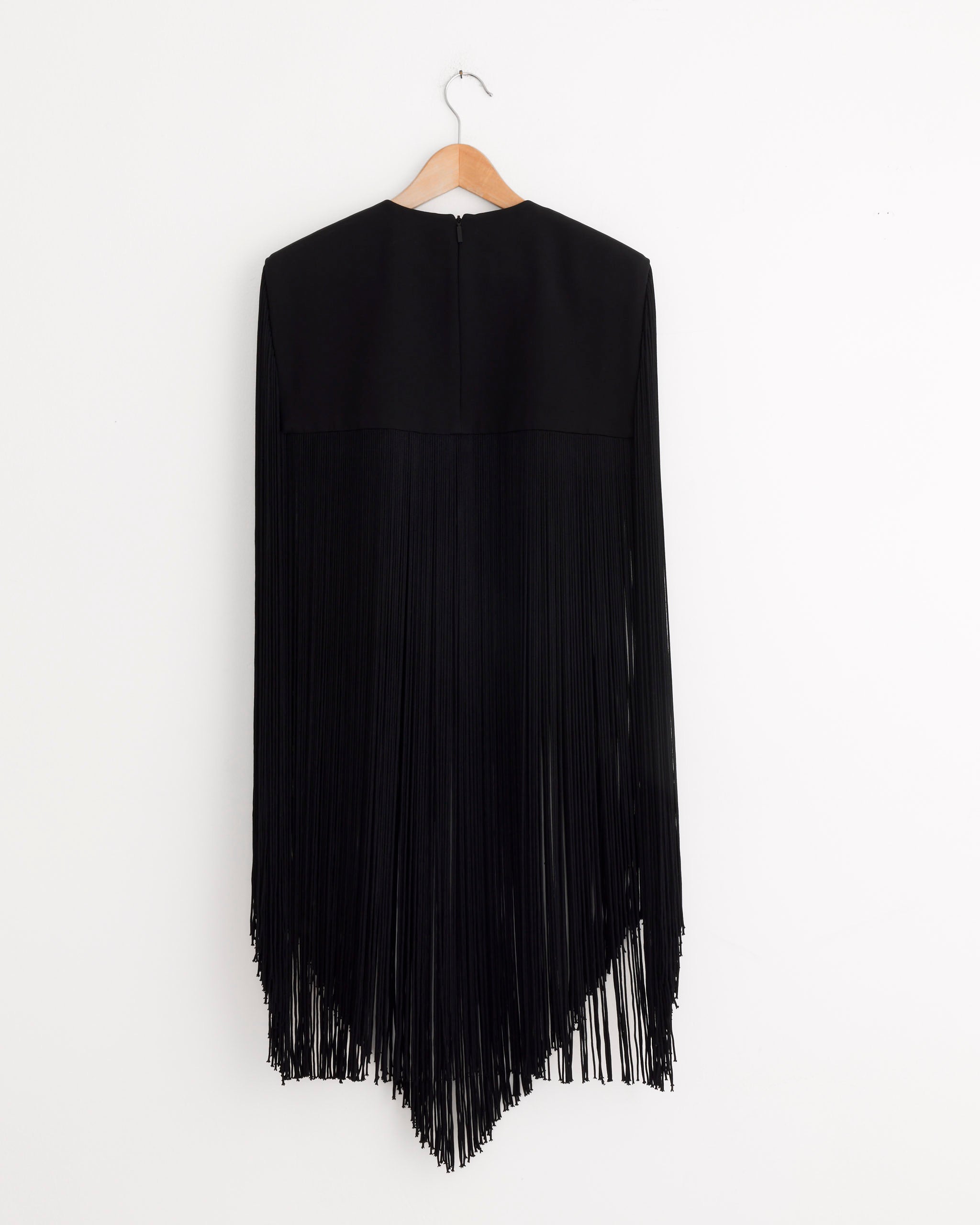 The Tibi 4-Ply Silk Fringe Cape in Black features long, vertical fringe and hangs elegantly from a wooden hanger against a white wall—an ideal evening cape for special occasions.