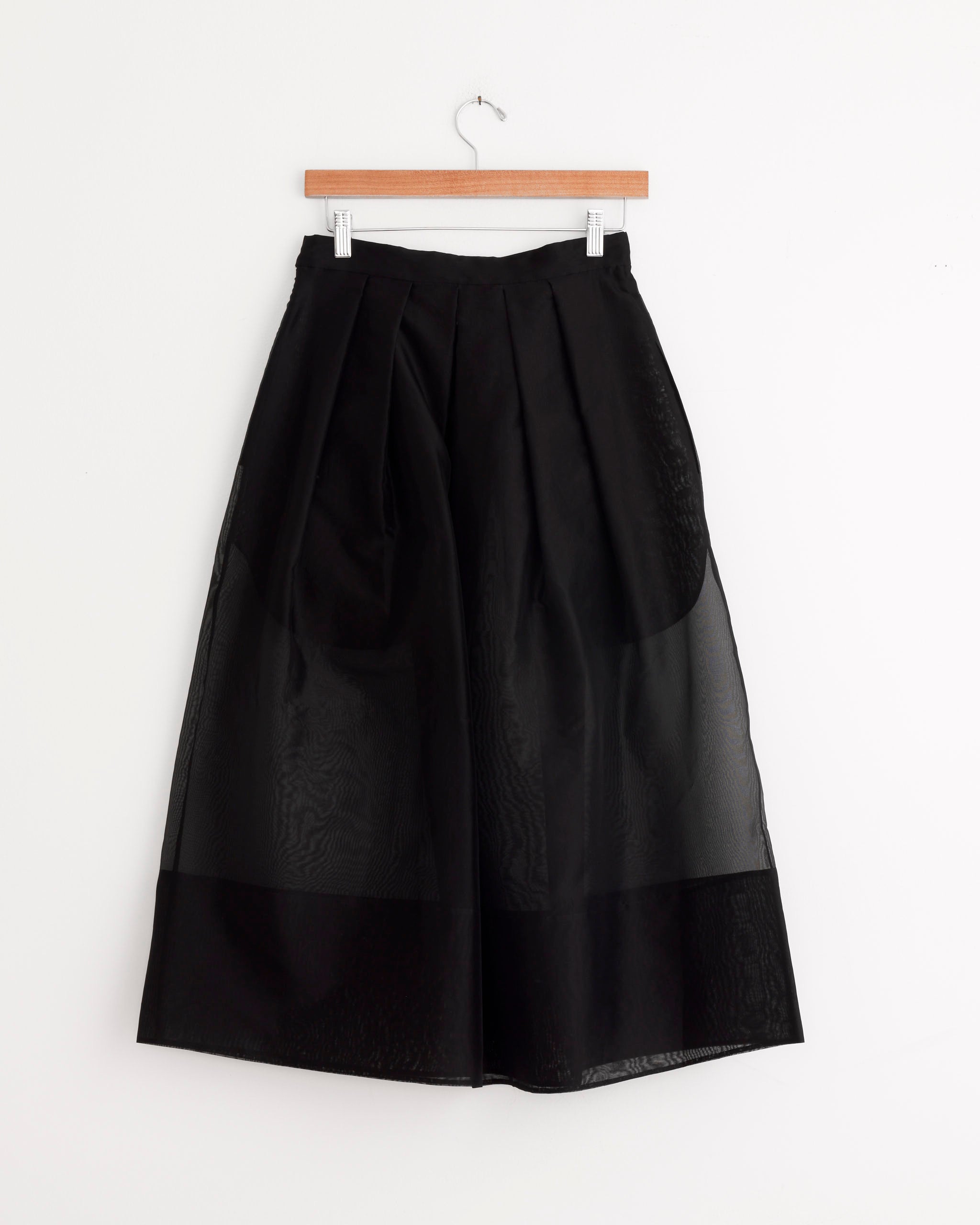 Tibi Organza Full Skirt in Black—a pleated, full midi skirt crafted from semi-sheer silk organza—hangs on a wooden hanger against a white background.