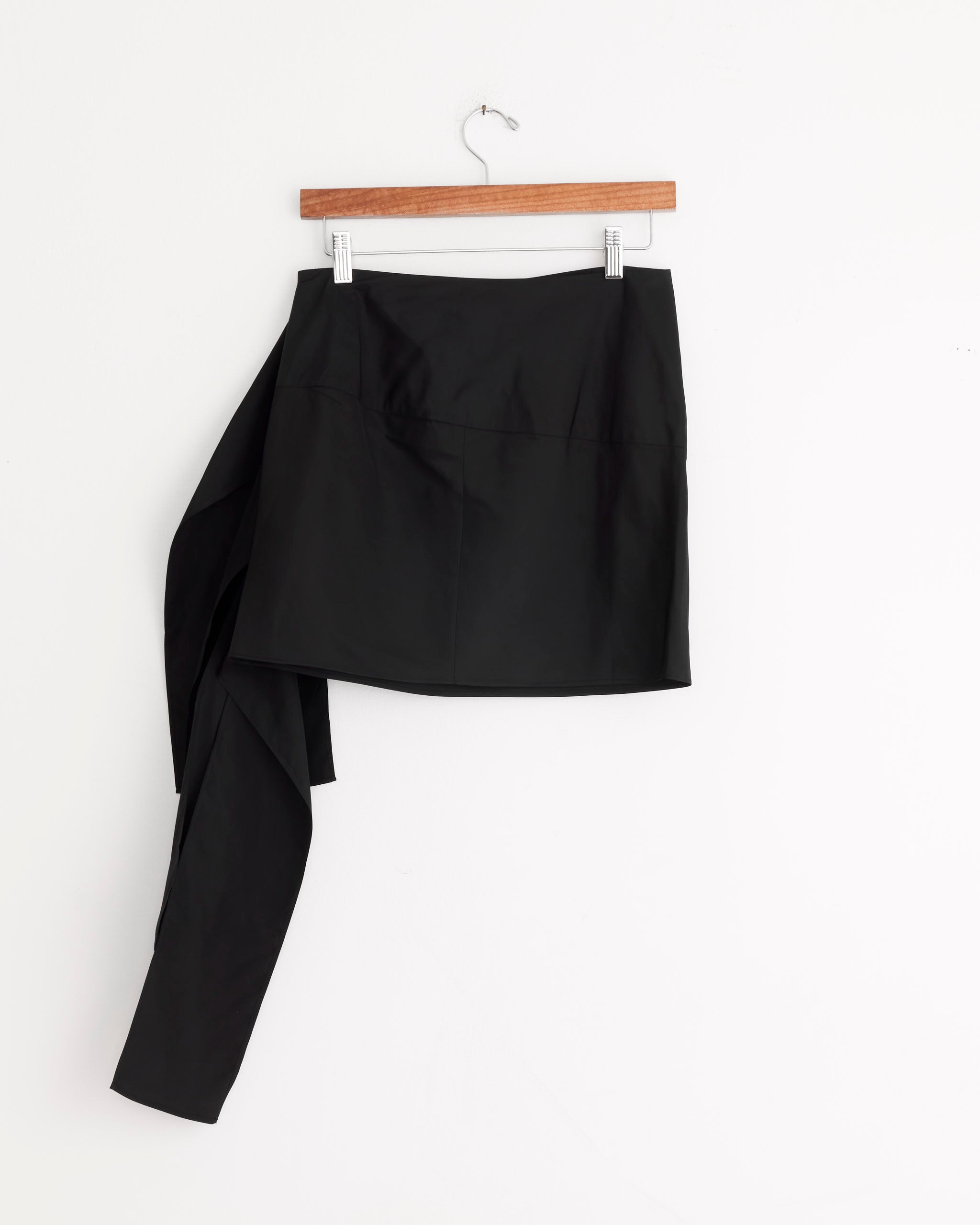 The Tibi Italian Sporty Ribbon Mini Skirt in Black, with a minimalist silhouette and an attached long, asymmetrical draped panel, is displayed on a wooden hanger against a white background.