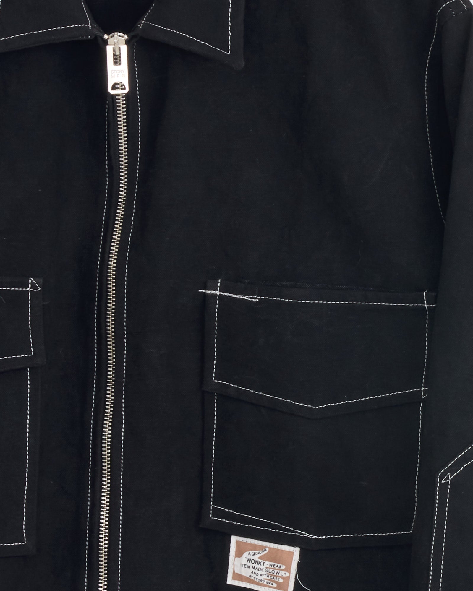 Close-up of the Story mfg 555 Jacket in Black Wonky Wear, made from organic cotton with white stitching, a silver zipper, two front flap pockets, and a small rectangular label on the bottom pocket.
