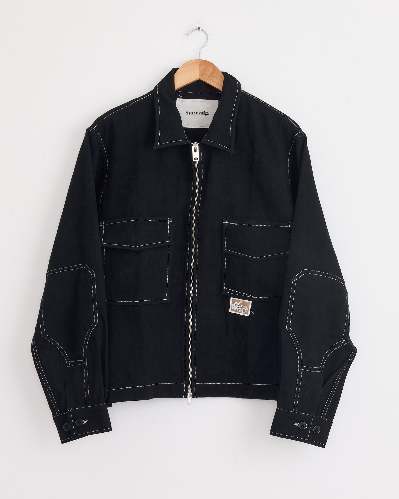 The 555 Jacket in Black Wonky Wear by Story mfg features white contrast stitching, large front pockets, and a small left pocket patch, shown on a wooden hanger against a white wall.