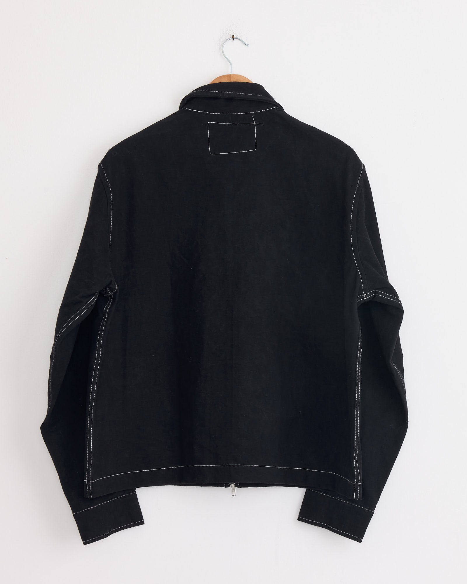 The Story mfg 555 Jacket in Black Wonky Wear, made from organic cotton with white contrast stitching, hangs on a wooden hanger against a plain white wall, shown from the back.