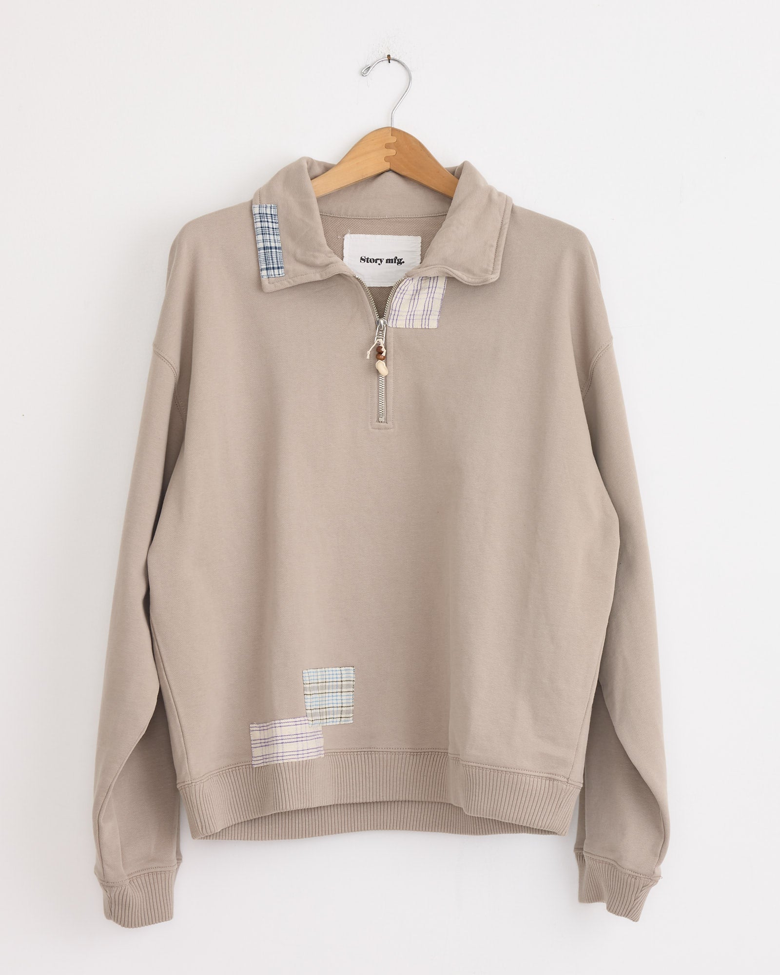 The Story mfg Geo Pullover in Grey Picnic, crafted from organic cotton, features a quarter-zip collar and blue plaid patchwork accents. Its vintage-inspired boxy fit hangs stylishly on a wooden hanger for timeless appeal.