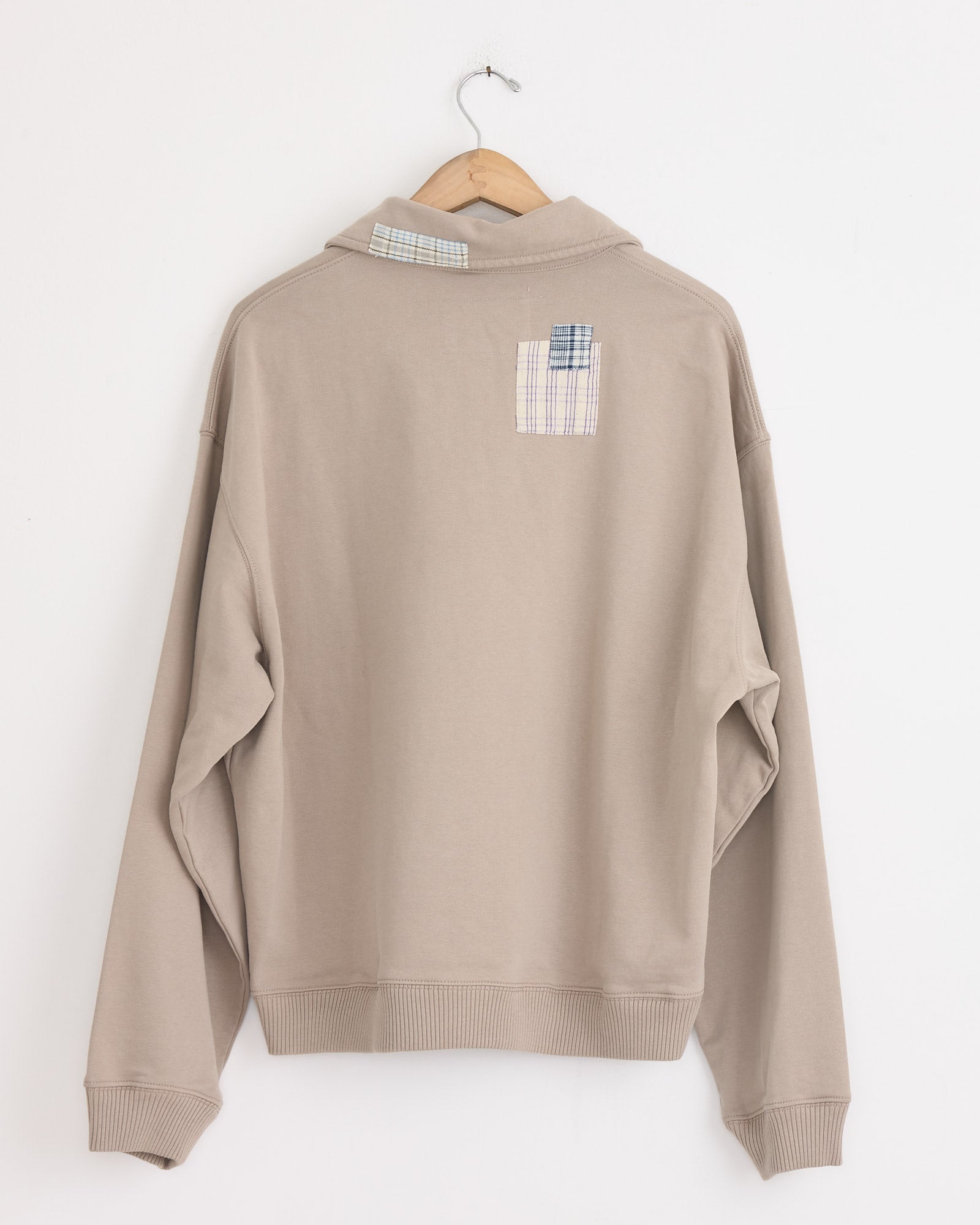 A grey organic cotton Geo Pullover by Story mfg with a vintage boxy fit hangs back-forward on a wooden hanger. Two small rectangular plaid patches on the upper and middle back add distinctive detail to this Grey Picnic edition.