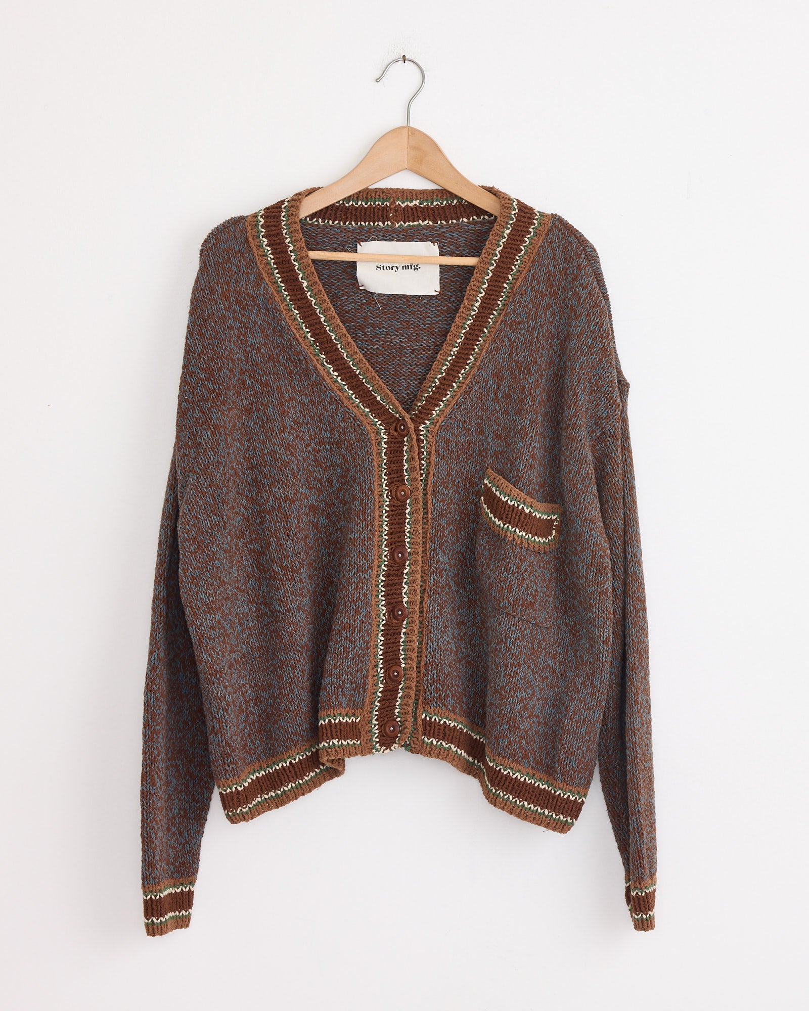The Bulb Cardigan in Twisted Dusk by Story mfg is a brown and blue hand-knit V-neck with front buttons, a small pocket, and decorative trim, crafted from organic cotton and displayed on a wooden hanger against a white wall.