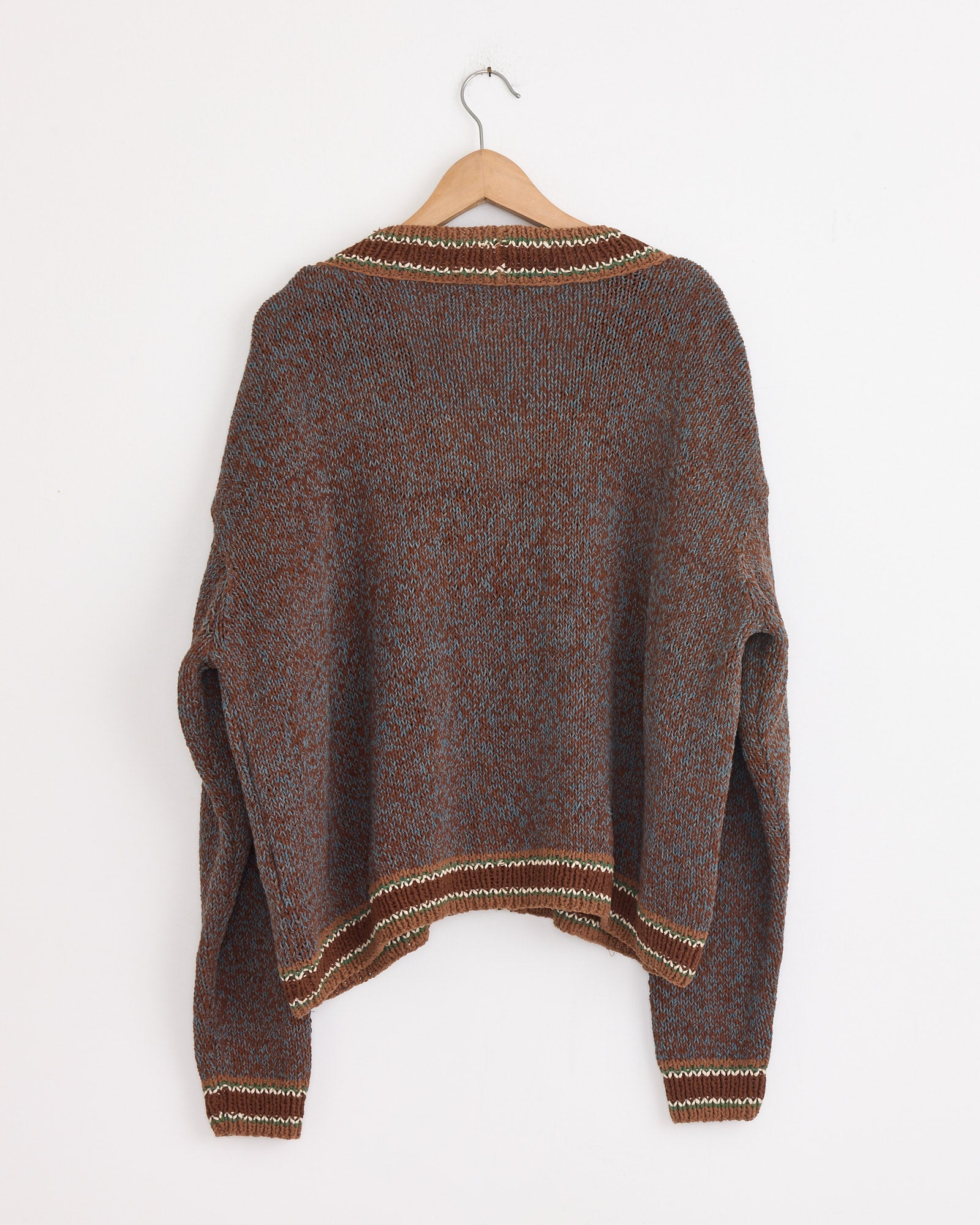 The Bulb Cardigan in Twisted Dusk by Story mfg is a hand-knit, organic cotton sweater in brown and gray with cream-striped collar, cuffs, and hem. Shown on a wooden hanger against a white wall with the back facing forward.