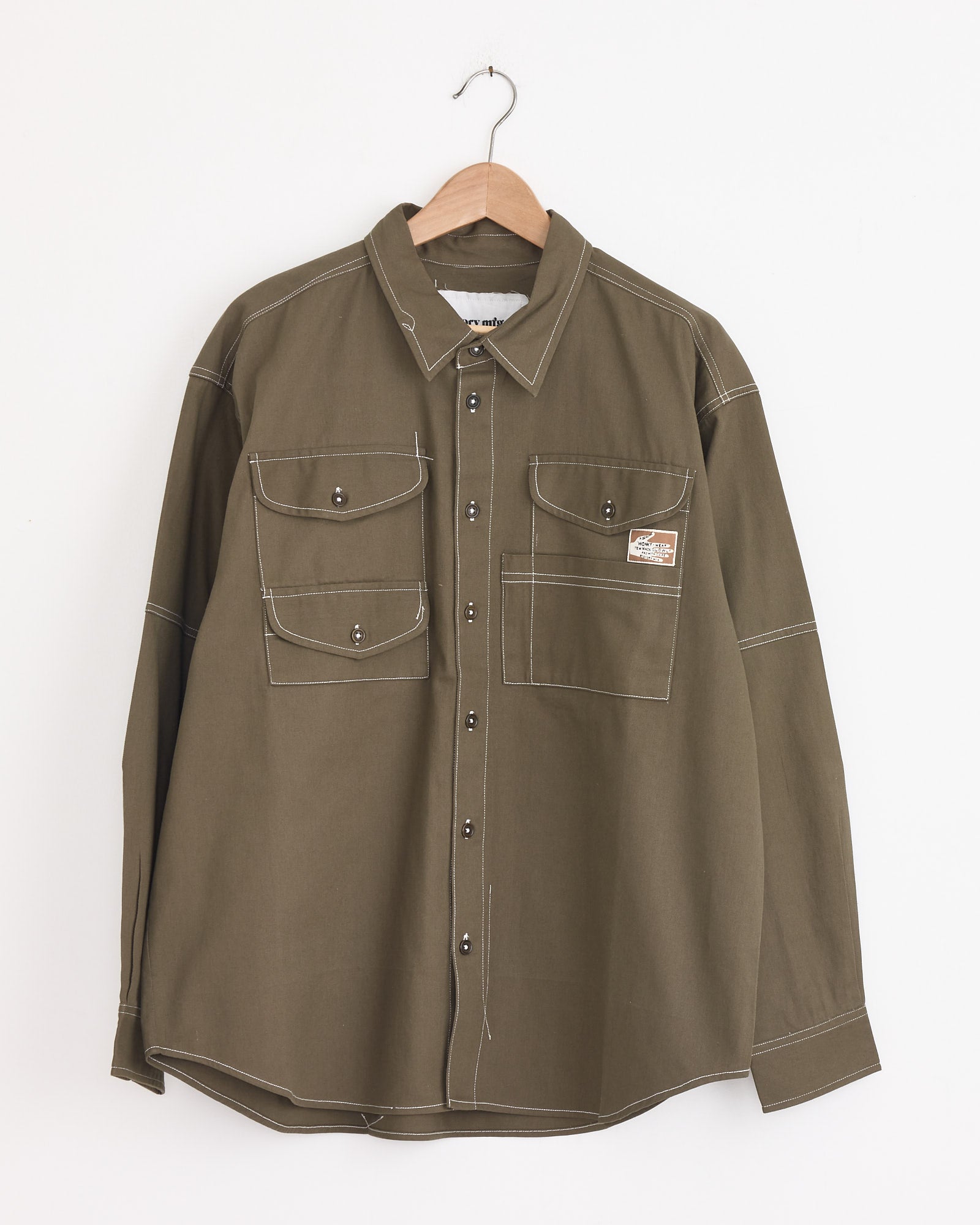 The Harvest Shirt in Khaki Wonky Wear by Story mfg features vintage workwear style, crafted from organic cotton with white stitching, two buttoned chest flap pockets, a logo patch on an open pocket, and is shown on a wooden hanger.