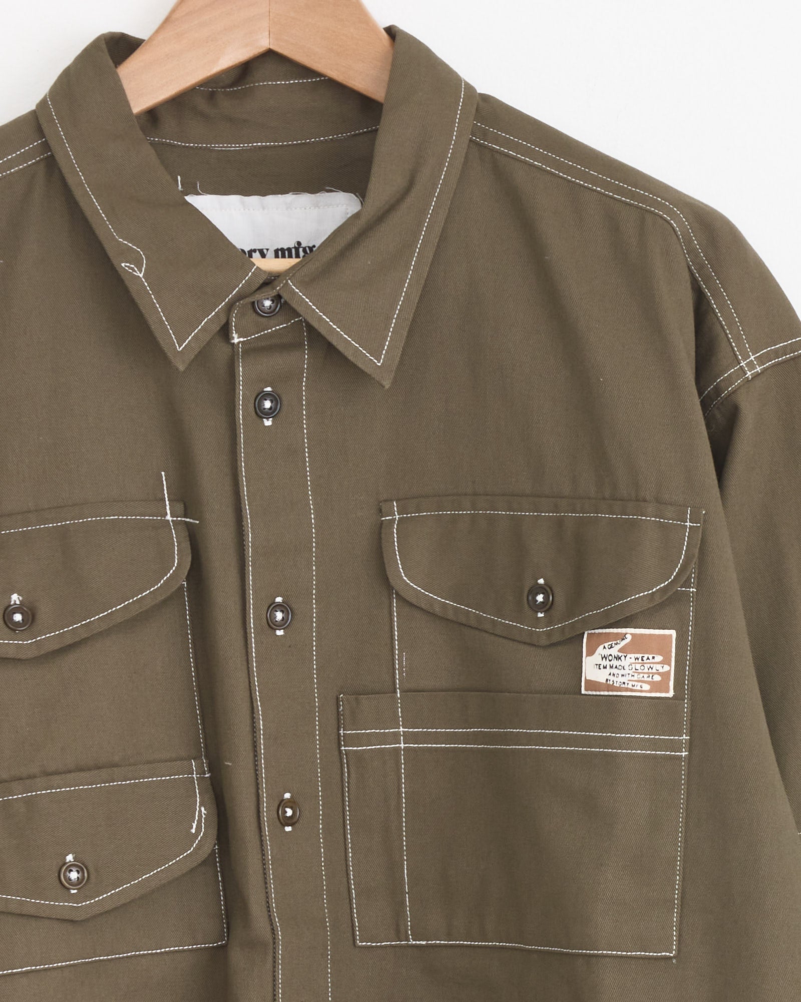 The Story mfg Harvest Shirt in Khaki Wonky Wear, made from organic cotton, features white stitching, two chest flap pockets with silver buttons, and a small patch on the right pocket. Shown on a wooden hanger against a white background.