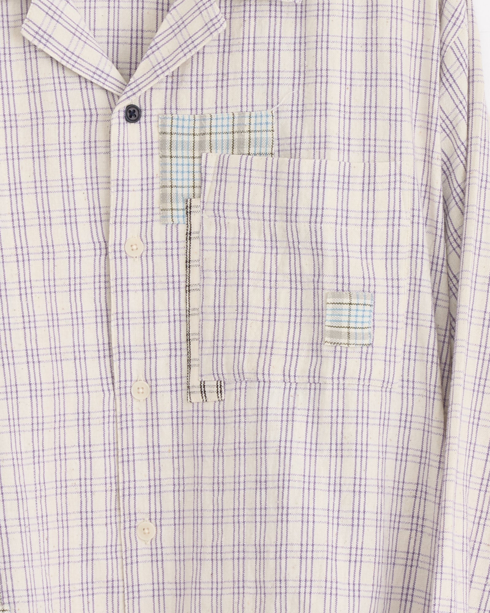 A close-up of the Story mfg Greetings Shirt in Purple Picnic, a light plaid organic cotton shirt with purple lines, featuring visible patchwork repairs in blue, beige, and black stitching by the chest pocket and buttons.