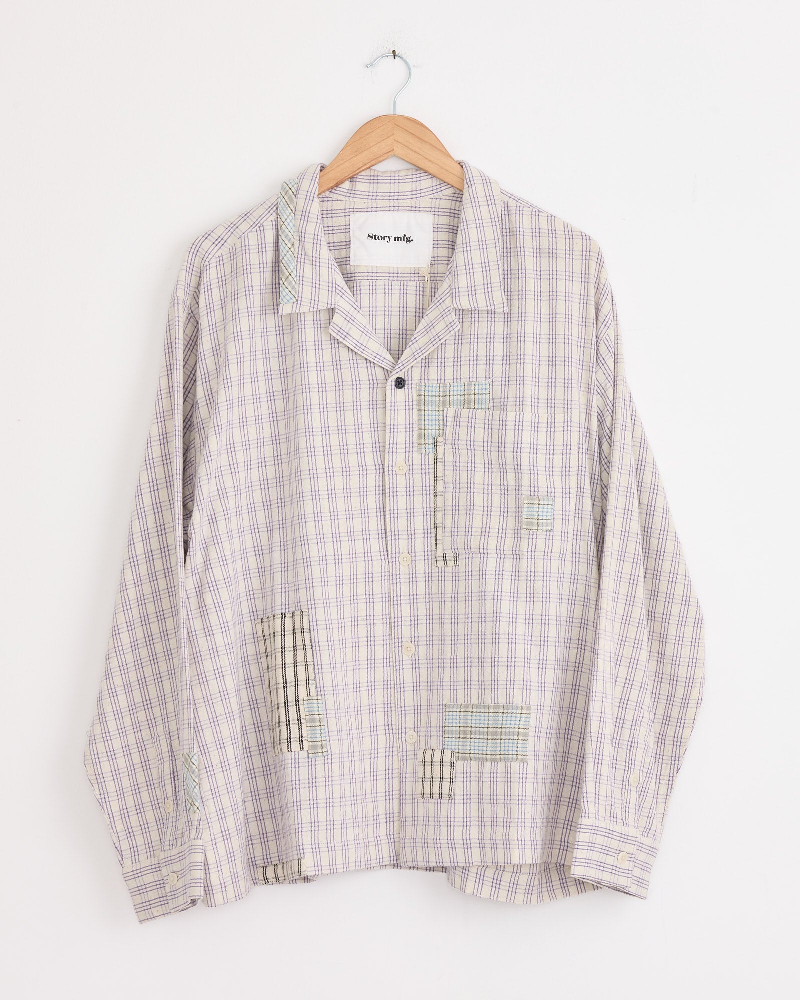The Story mfg Greetings Shirt in Purple Picnic is a long-sleeved, boxy-fit, handwoven cotton shirt with patchwork details, a single chest pocket, and a loose silhouette, shown on a wooden hanger against a white wall.