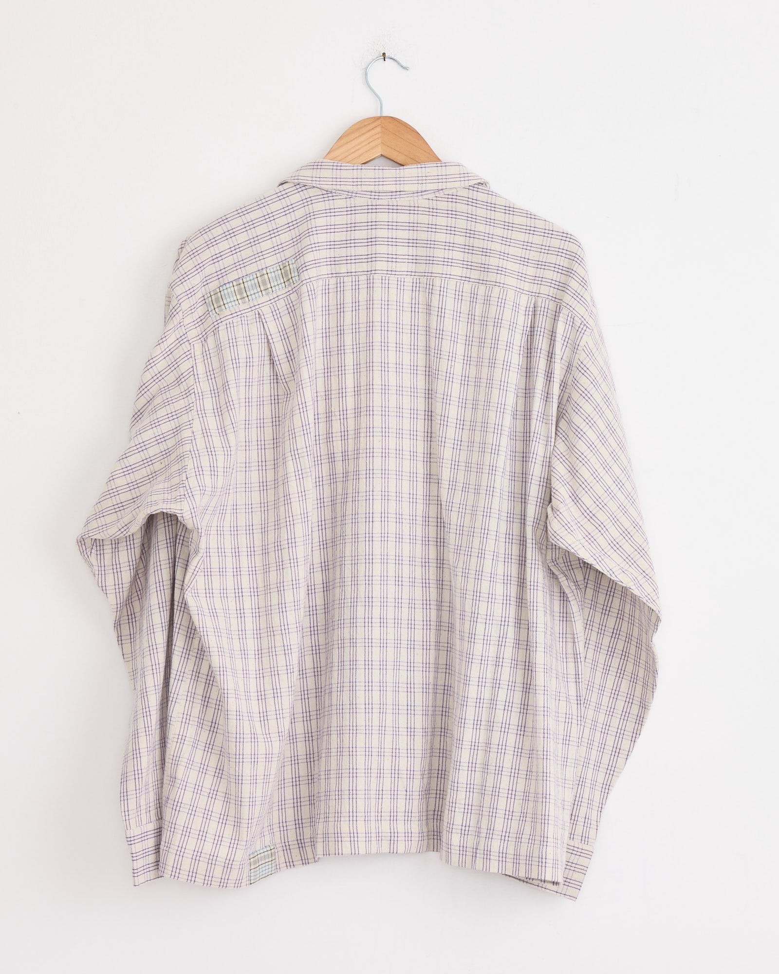 The Story mfg Greetings Shirt in Purple Picnic is a loose, boxy plaid shirt with blue and pink stripes, shown from the back on a wooden hanger against a plain white wall.