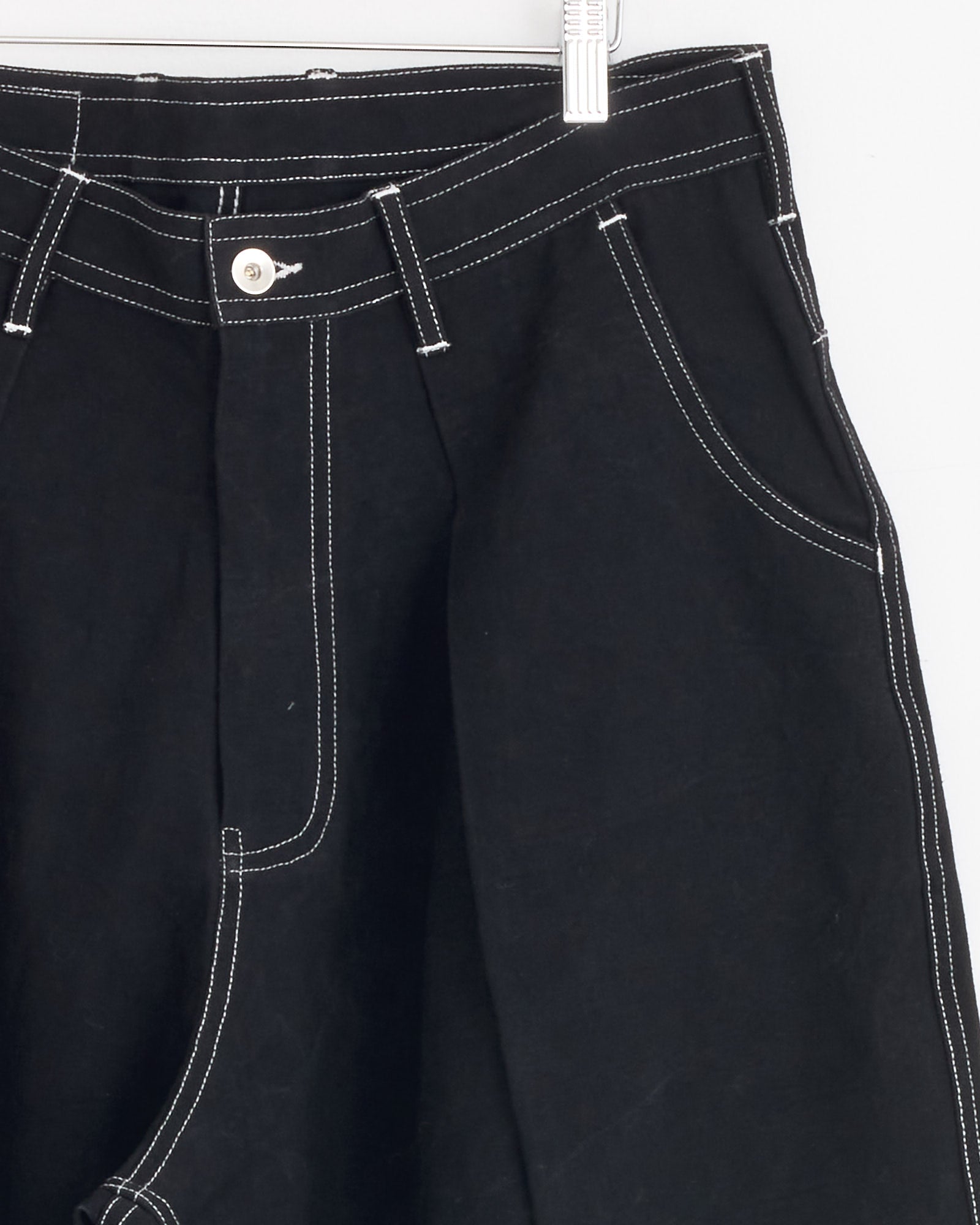 Close-up of Story mfg Lush Pant in Black Wonky Wear, featuring workwear style with white contrast stitching, silver button, belt loops, pockets, and zipper fly, hanging on a white hanger against a white background.
