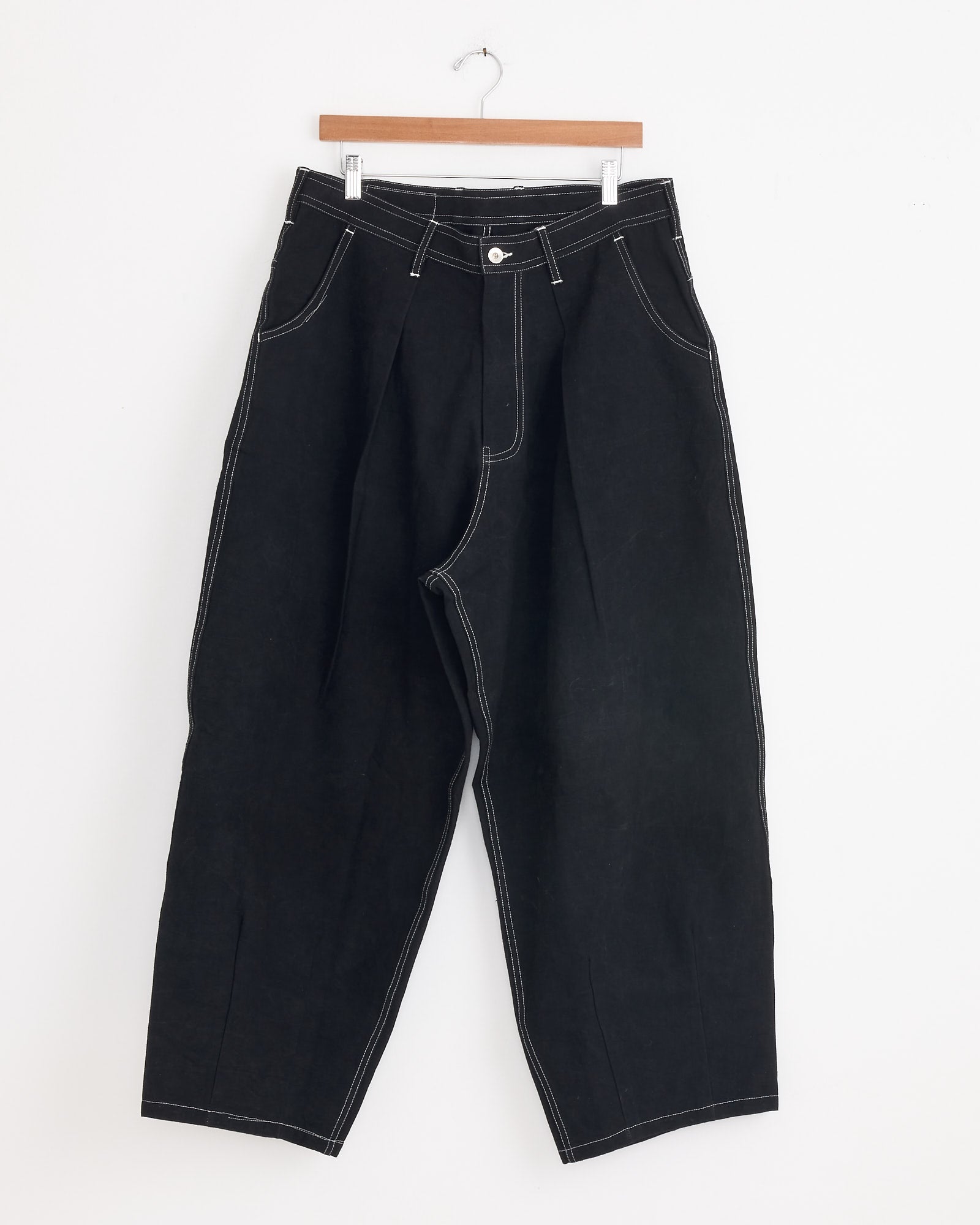 The Lush Pant in Black Wonky Wear by Story mfg is a pair of loose-fitting black workwear trousers with white contrast stitching, pleated waist, and a button fly, displayed on a wooden hanger against a white background.