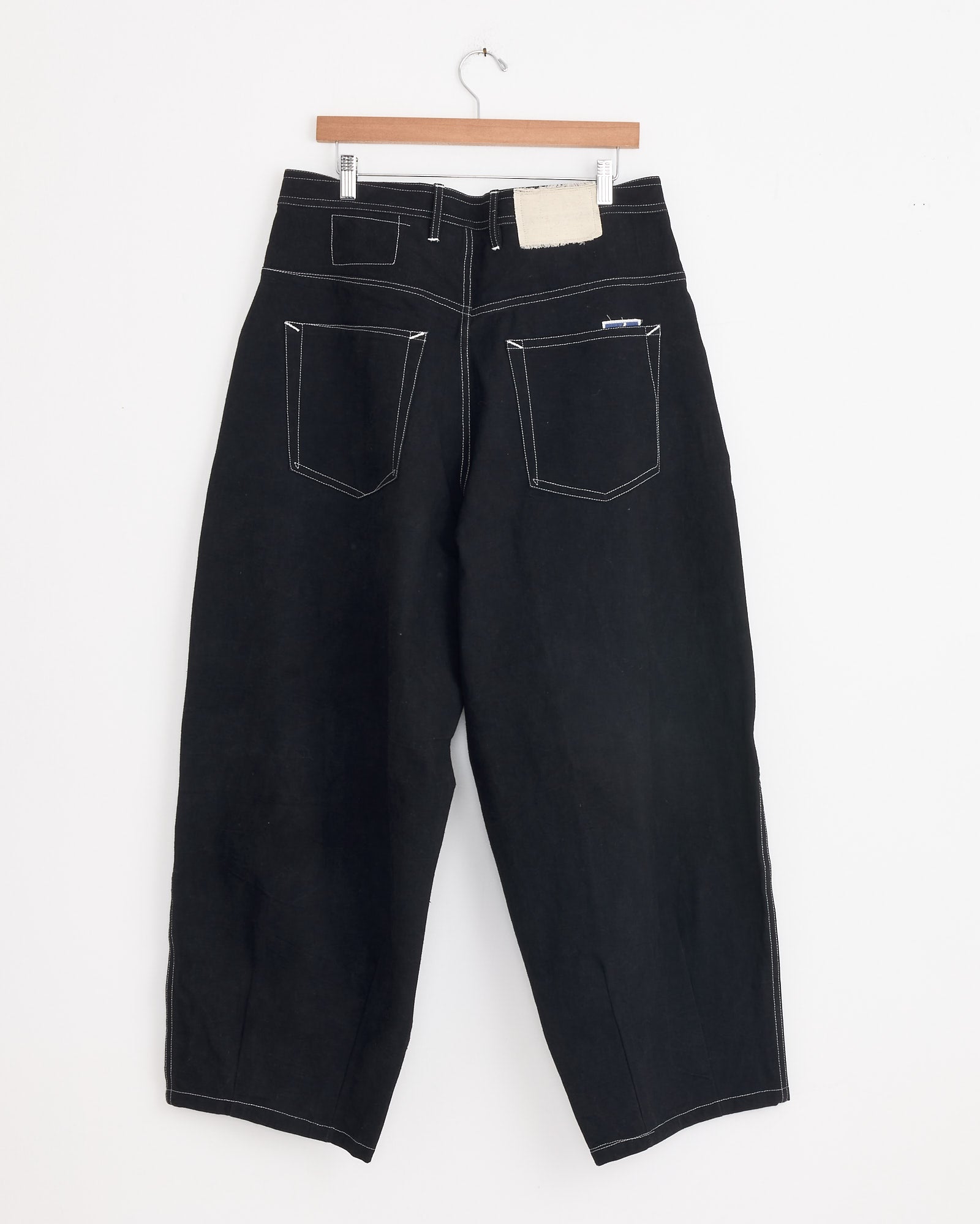 The Lush Pant in Black Wonky Wear by Story mfg features wide legs, white stitching, two large back pockets, and a white patch near the waistband, shown hanging on a wooden hanger against a white background.