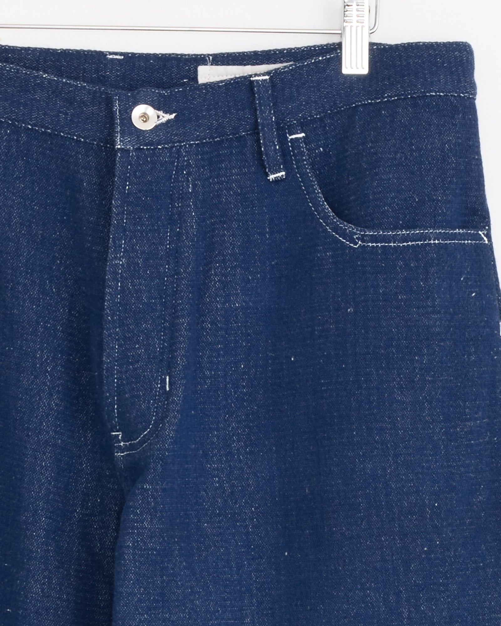 Close-up of the upper part of the Poetry Jean in Selvedge Denim by Story mfg, featuring blue denim, white stitching, a metal button, belt loops, and a front pocket. This vintage-inspired piece hangs on a white hanger against a plain background.