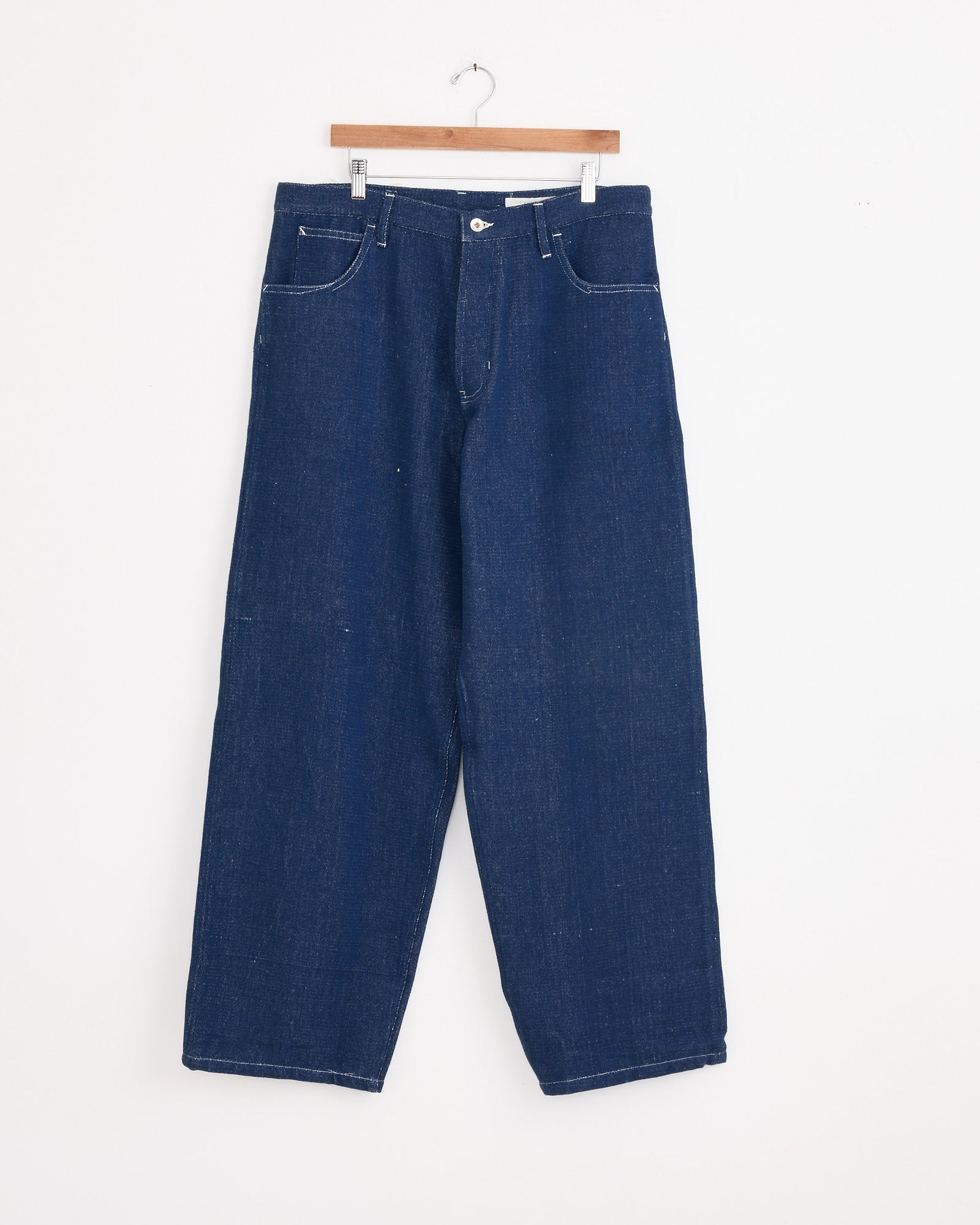 The Poetry Jean in Selvedge Denim by Story mfg is a dark blue straight-leg jean with white stitching, front button and zipper, and two pockets, crafted from handwoven selvedge denim and displayed on a wooden hanger against a white wall.
