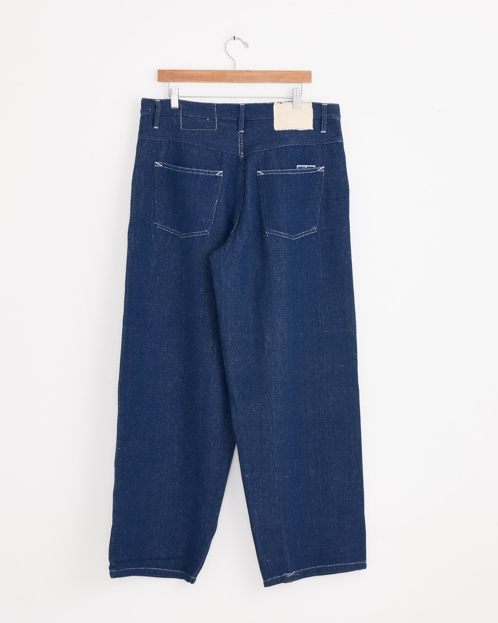 A pair of Story mfg Poetry Jean in Selvedge Denim hangs on a wooden hanger against a white wall, shown from the back with two rear pockets and visible stitching, evoking classic vintage workwear style.