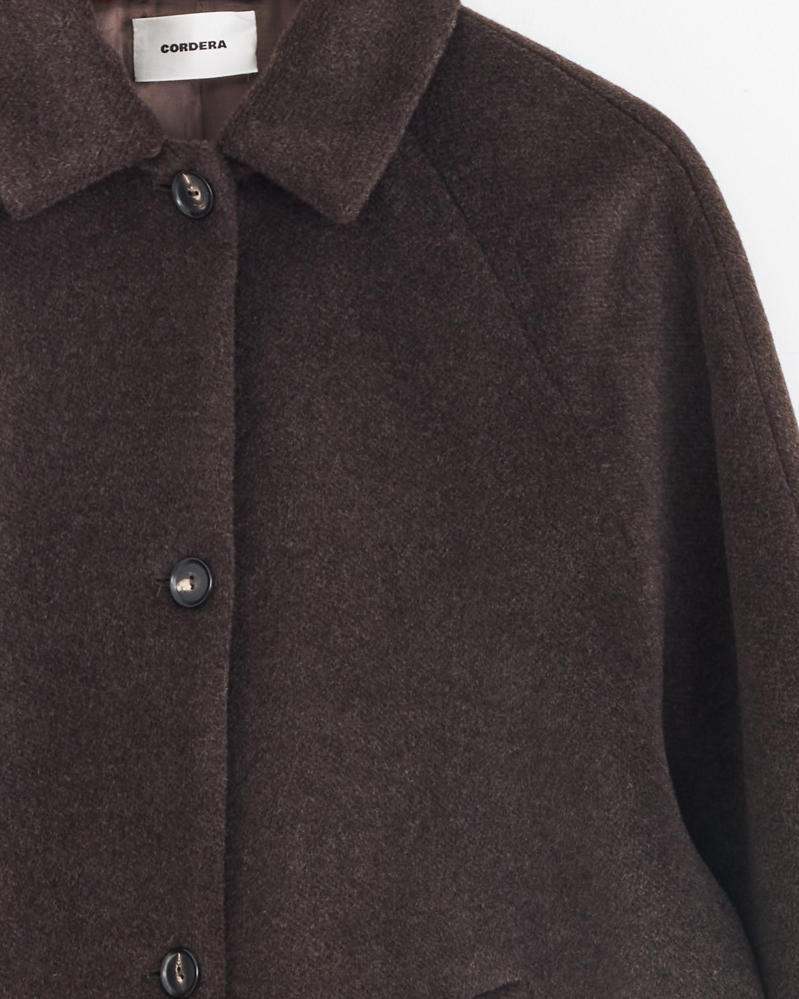 Close-up of Cordera’s Single Breasted Wool Coat in Mocca, featuring black buttons and a spread collar. The Cordera label is visible inside the collar. Only the upper section and part of a sleeve appear against a plain background.