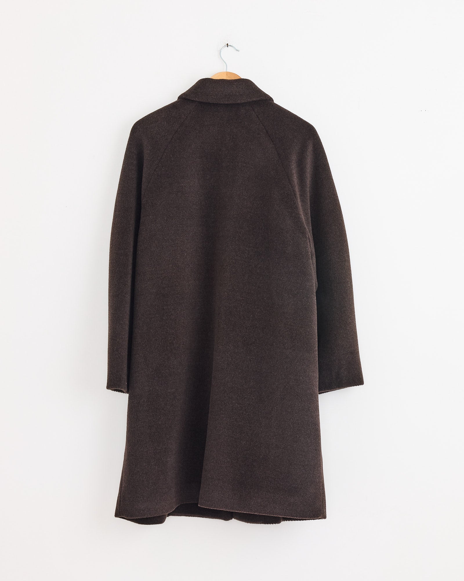 The Cordera Single Breasted Wool Coat in Mocca, shown from the back, is oversized and dark brown, hanging on a wooden hanger against a plain white wall.