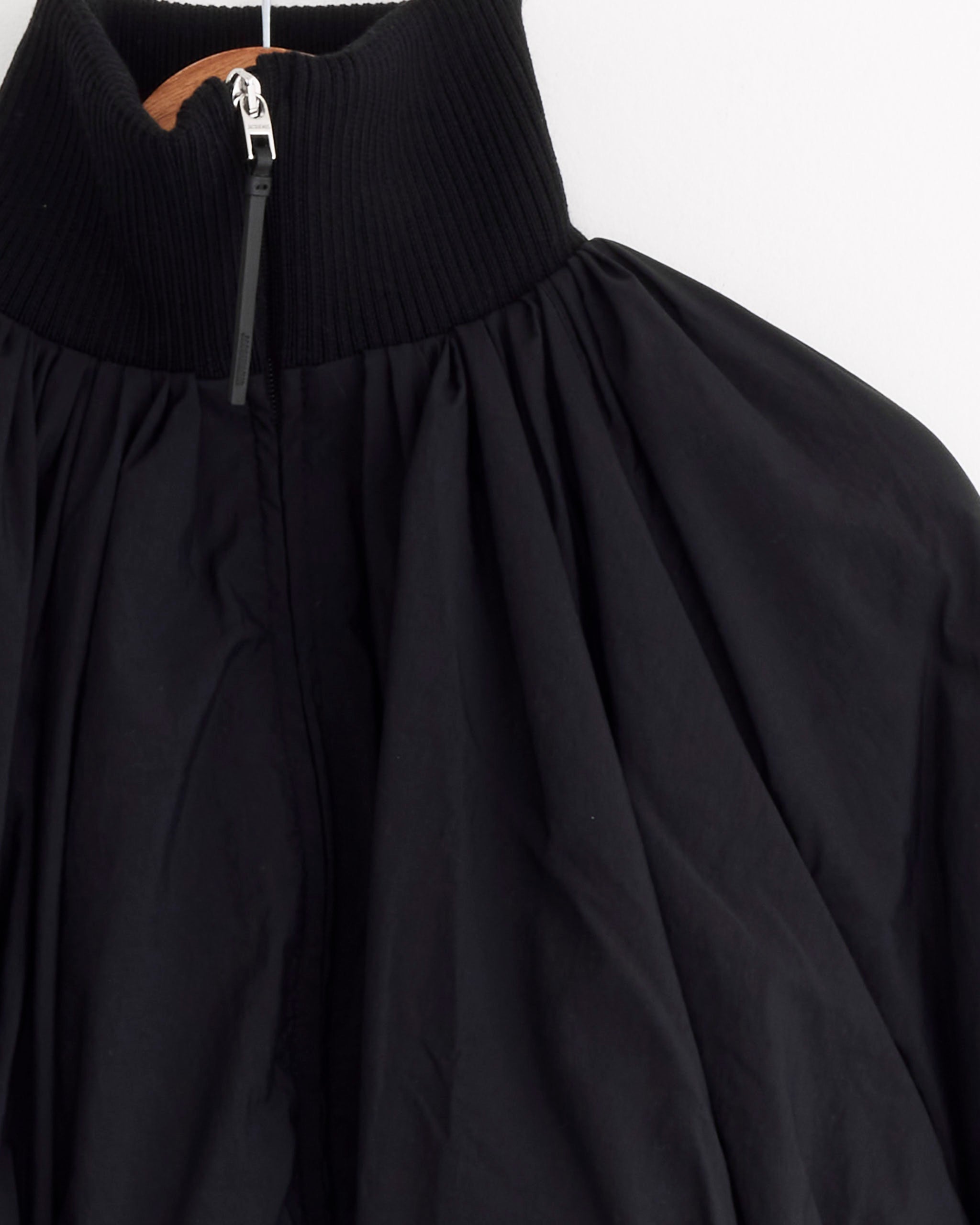 A close-up of the Jacquemus Le Bomber Pallone in black, featuring a ribbed high collar, zipper, and draped nylon fabric, hanging on a wooden hanger against a white background.