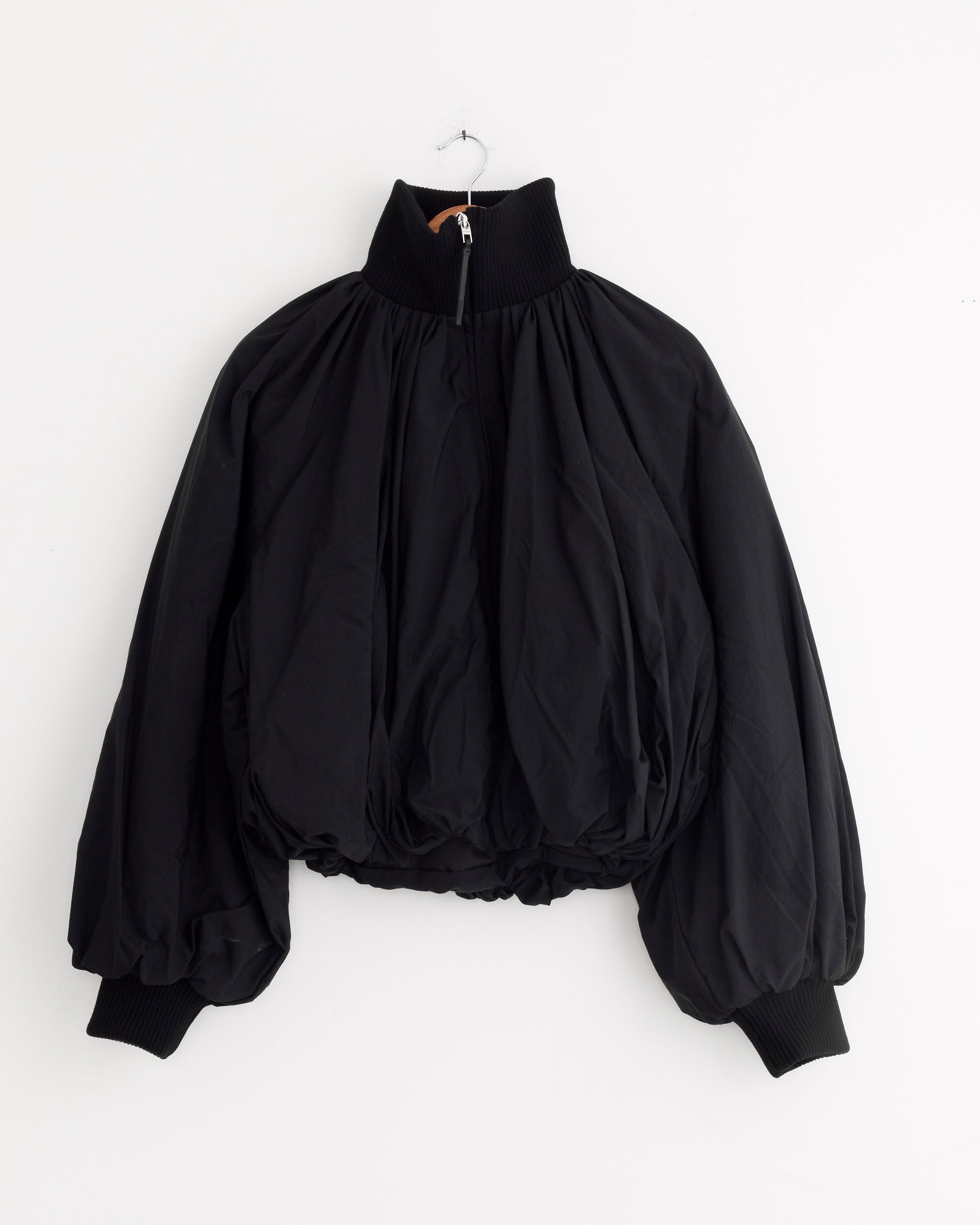 The Jacquemus Le Bomber Pallone in Black, crafted from draped nylon with puffy gathered fabric, ribbed cuffs, a high ribbed collar, and long sleeves, hangs on a white wall by a hanger.