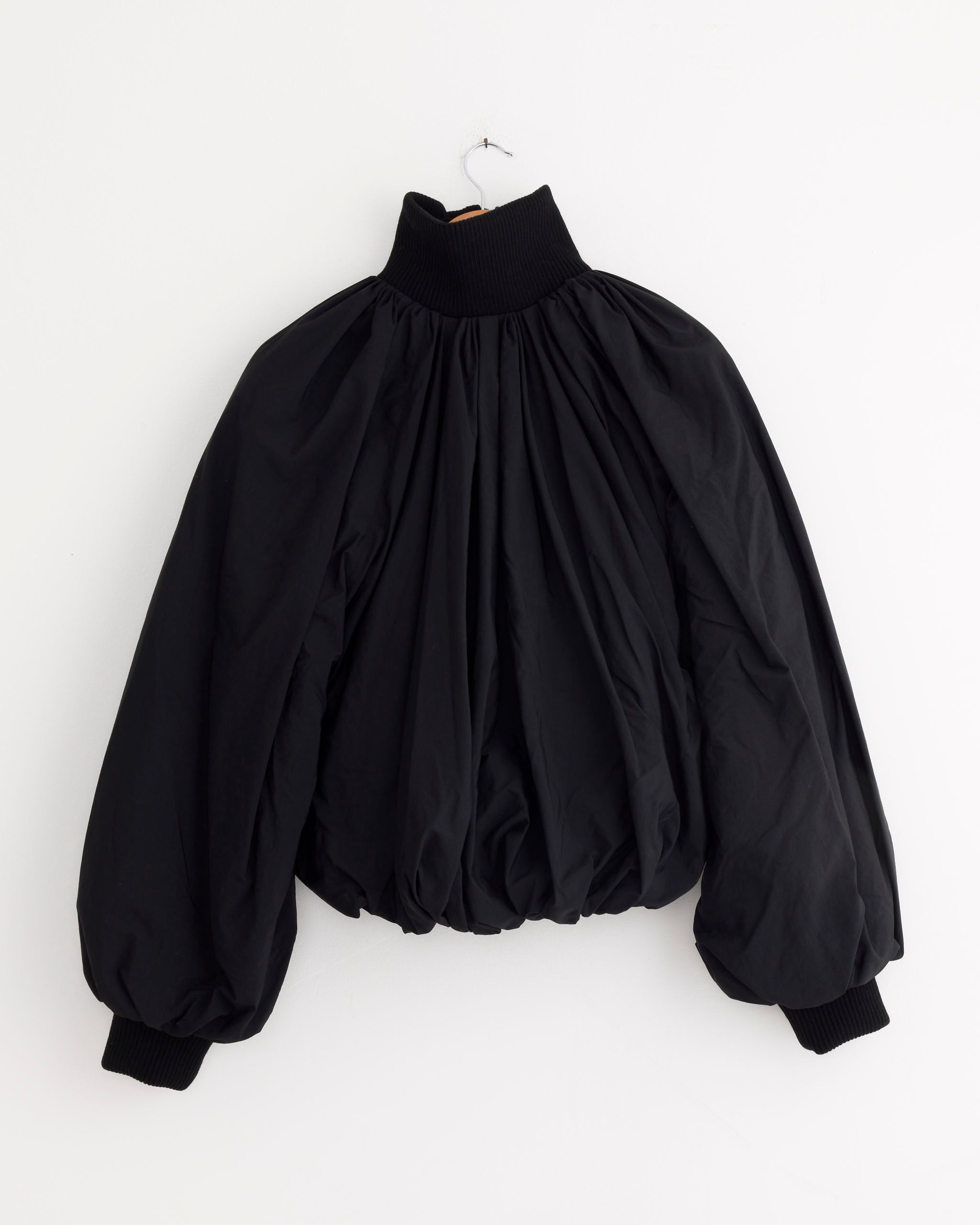 The Jacquemus Le Bomber Pallone in Black, a puffy bomber jacket with voluminous balloon sleeves, high ribbed collar, and ribbed cuffs in draped nylon, hangs on a simple hook against a plain white background.