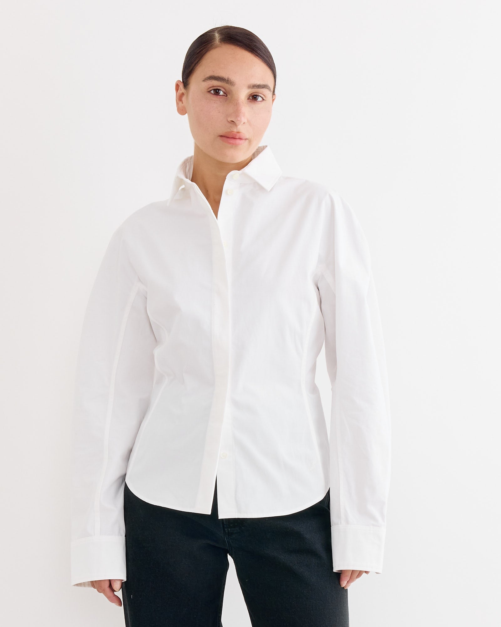 A person with dark hair in a neat bun wears the Jacquemus La Chemise Ovalo in White, paired with black pants, standing against a plain white background and looking at the camera.