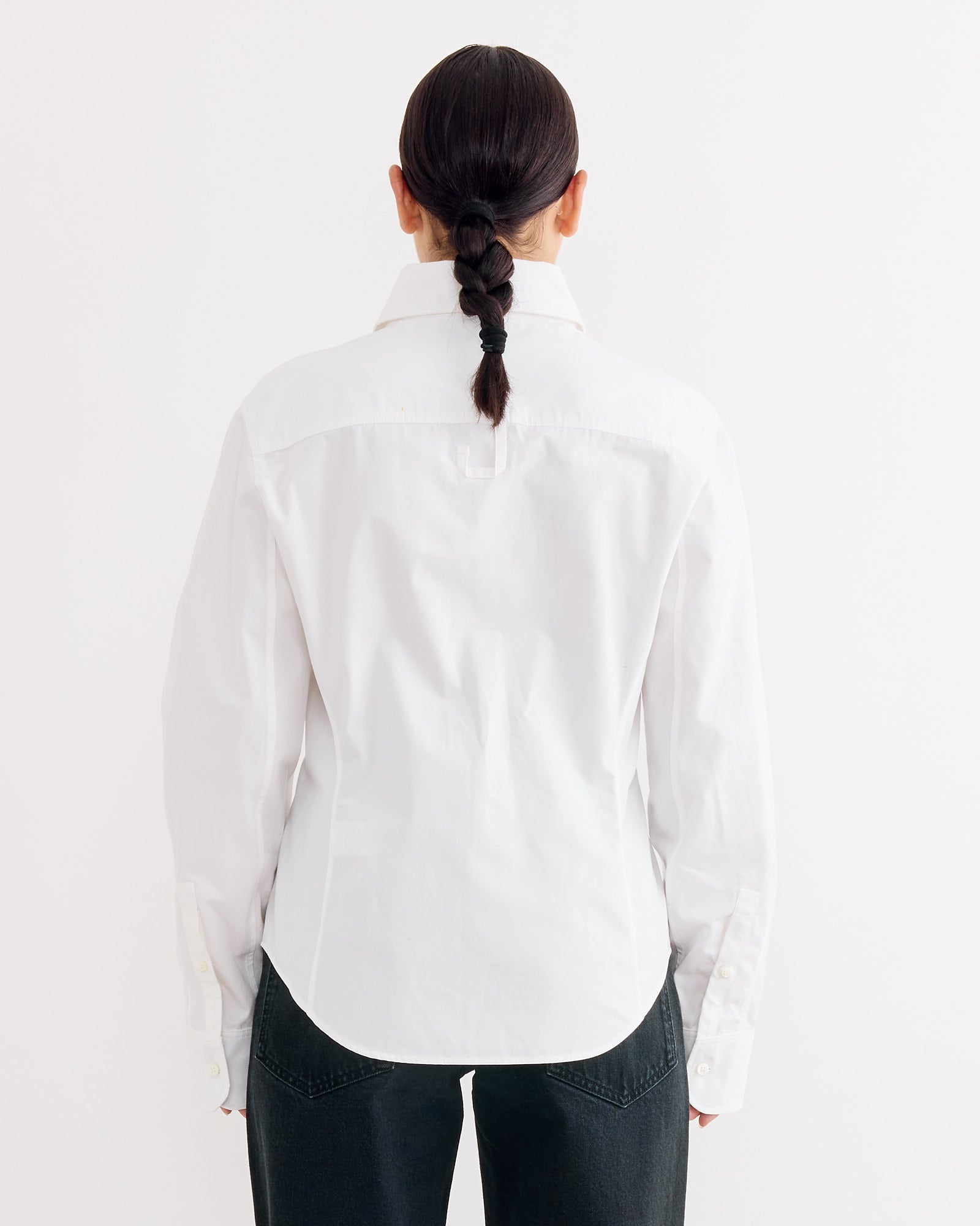 A person with dark hair in a single braid is seen from behind, wearing the Jacquemus La Chemise Ovalo in White, paired with black pants, standing against a plain white background.