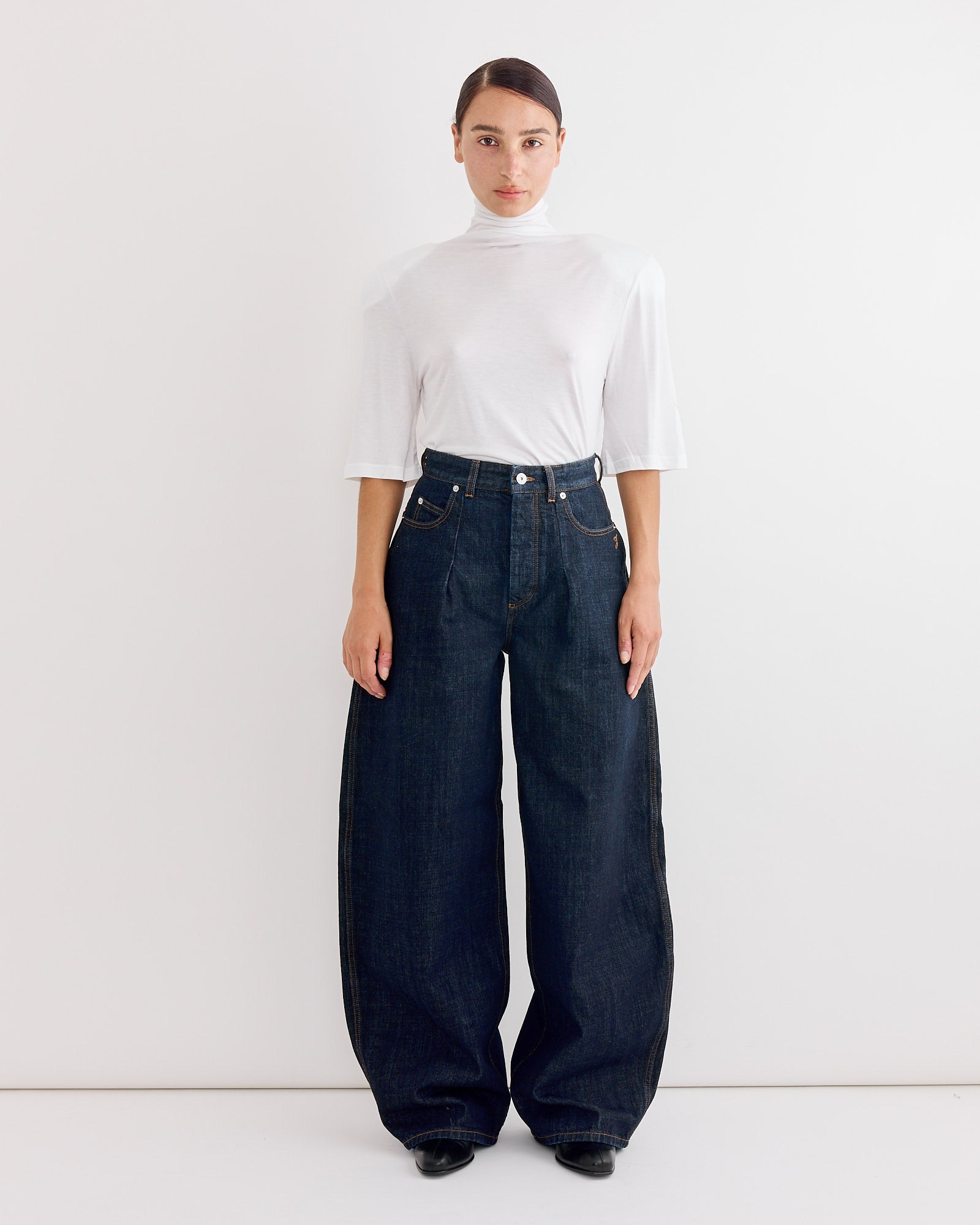 A woman stands against a white background wearing the Jacquemus Le De-Nimes Ovalo Pant in Dark Navy, paired with a white turtleneck and black shoes, highlighting the flattering high-waisted, wide-leg silhouette of this classic style.