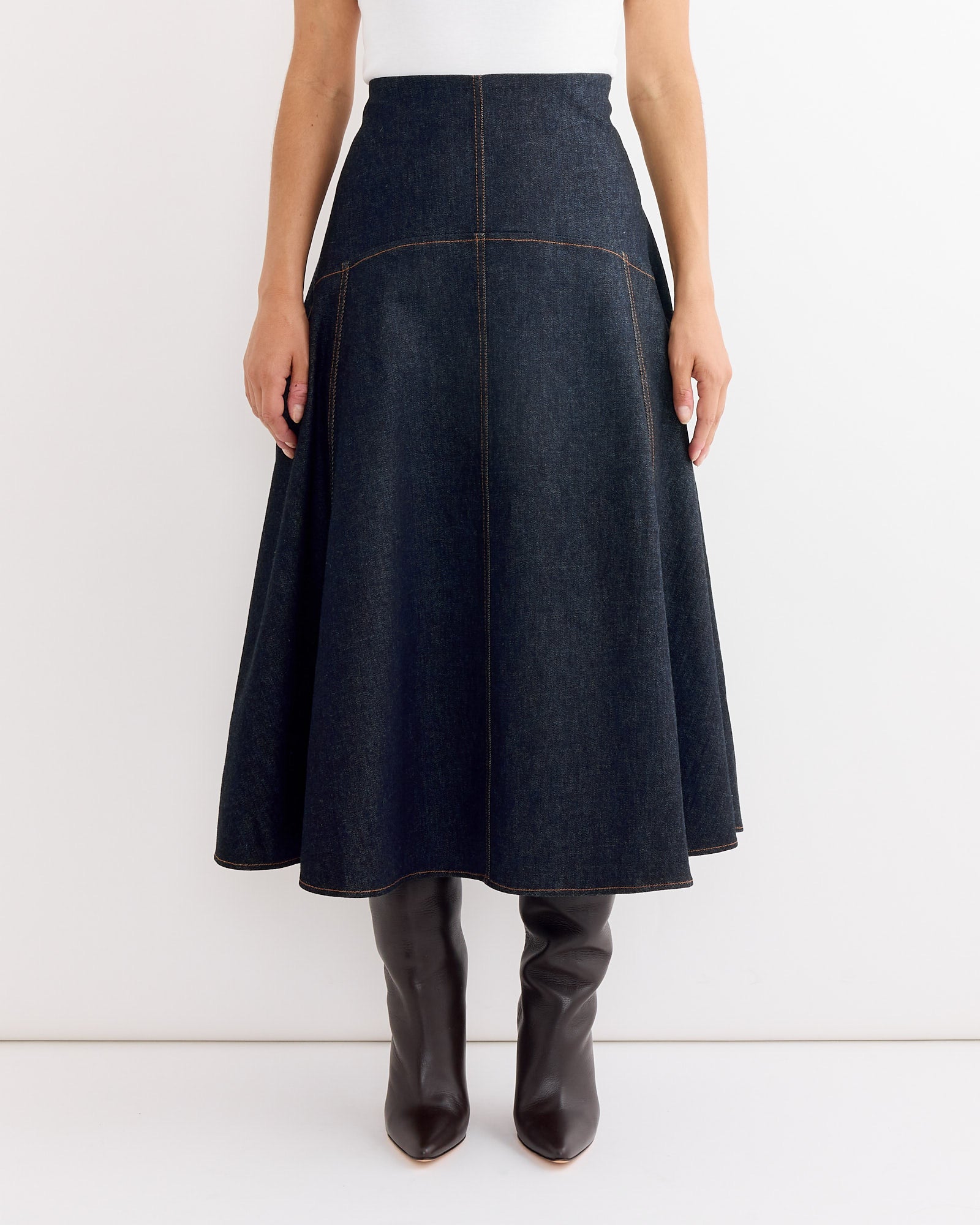 A person wears the Jacquemus La Jupe Triangle De-Nimes Skirt in Dark Navy with a white top and dark brown knee-high boots, standing against a plain white background. For fit questions, our online chat can help.