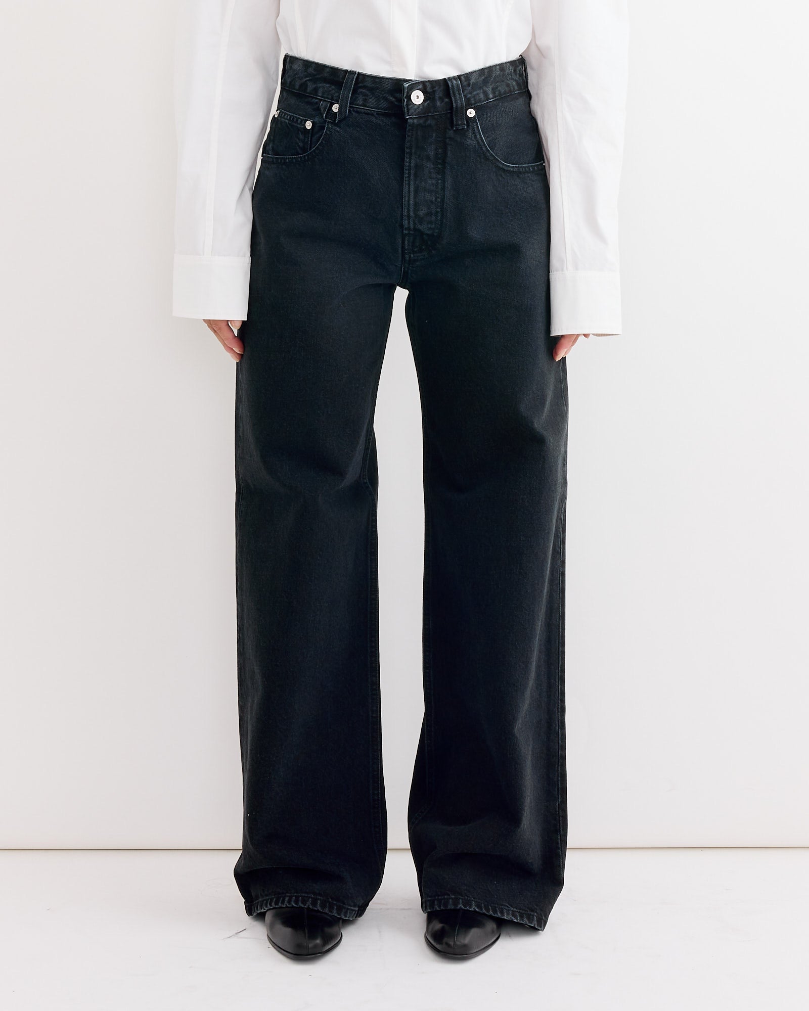 A model showcases the Jacquemus Le De-Nimes Large Pant in Black—high-waisted, wide-leg jeans—paired with a white long-sleeve shirt, photographed from shoulders down against a plain white background to highlight fit and sizing.