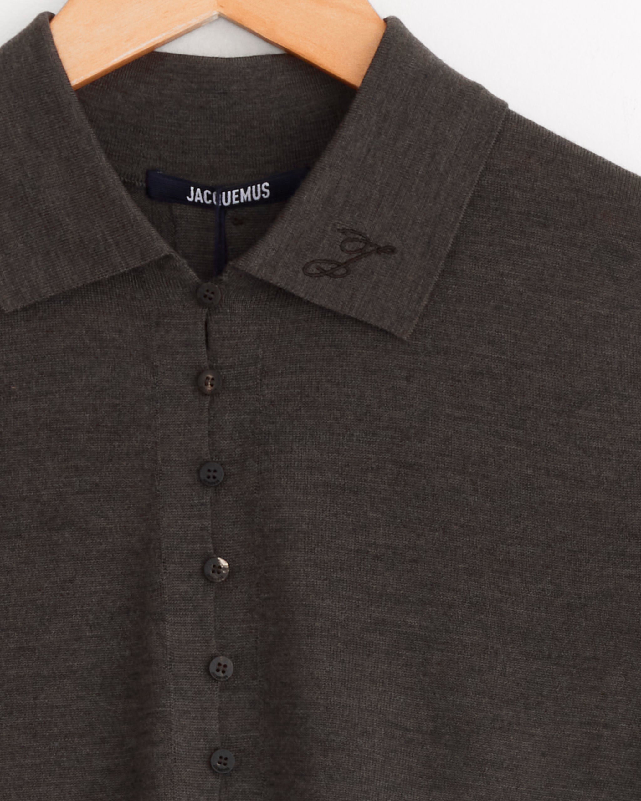 A dark brown Jacquemus Le Polo Merino Top with front buttons hangs on a wooden hanger. Embroidered details accent the collar, and a fitted merino wool design reveals the JACQUEMUS label inside.