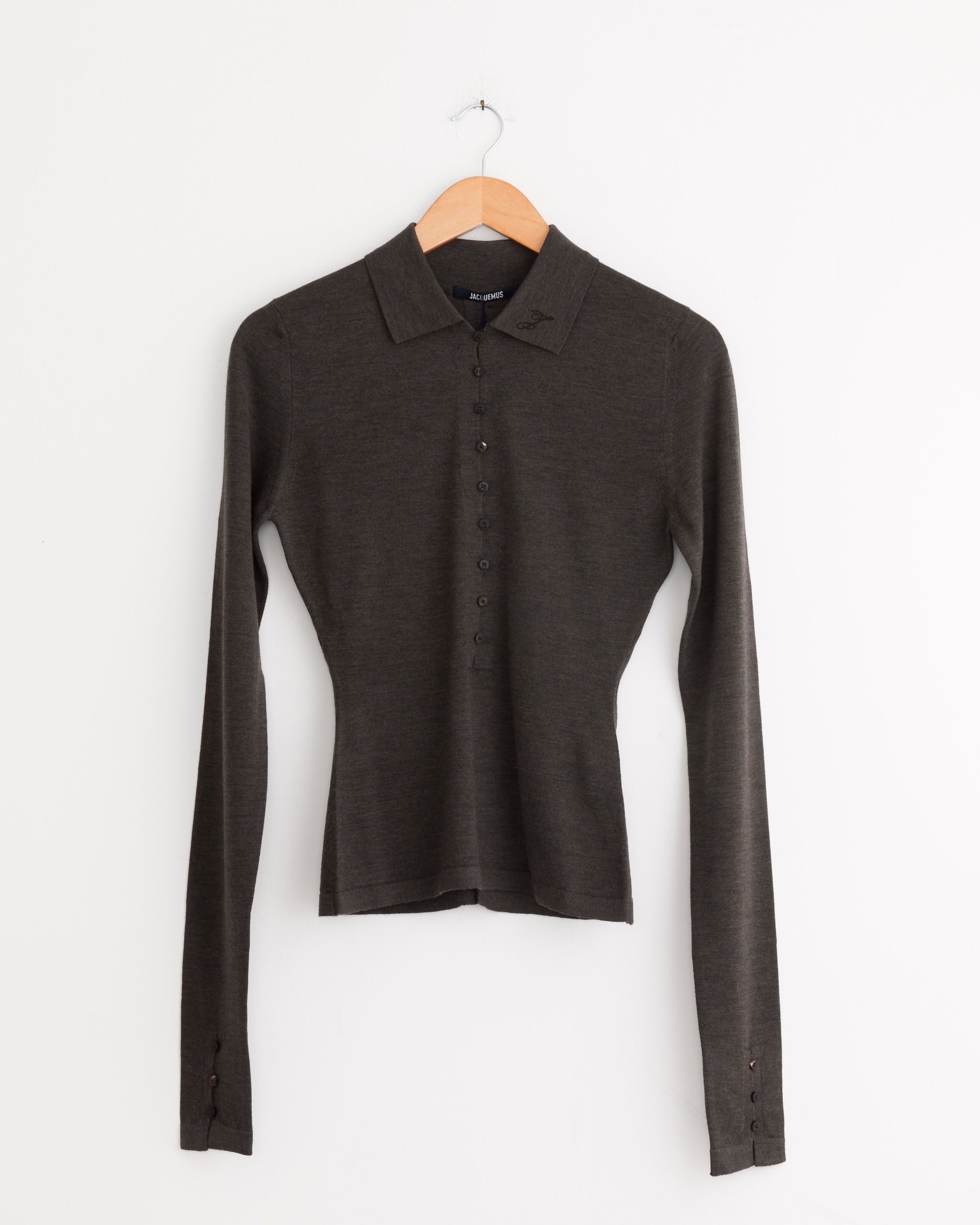The Jacquemus Le Polo Merino Top in Dark Brown, a long-sleeve collared button-up, is displayed on a wooden hanger against a white wall.