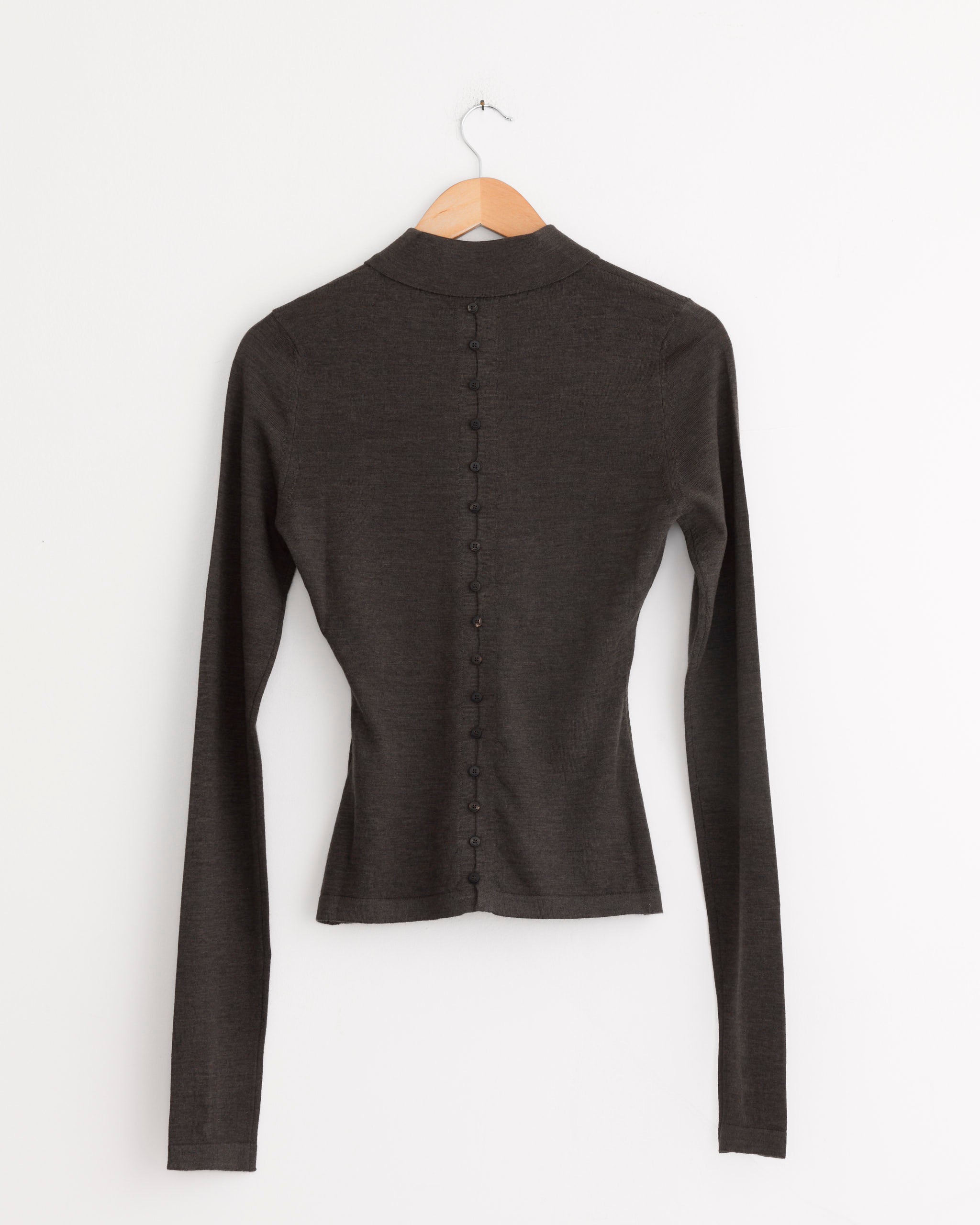 The Jacquemus Le Polo Merino Top in dark brown, a long-sleeved fitted piece, is displayed on a wooden hanger against a white wall, shown from the back with small buttons running down the center.