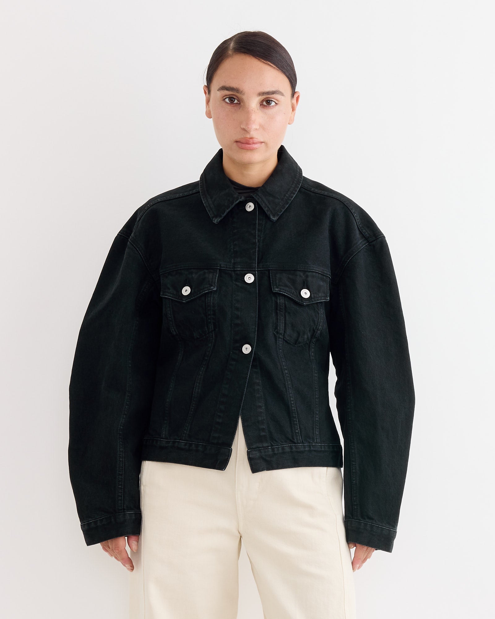 A person stands against a white background, wearing the Jacquemus La Veste De-Nimes Jacket in Black with silver buttons, classic collar, and front pockets, paired with light beige pants. For fit questions, try our online chat.