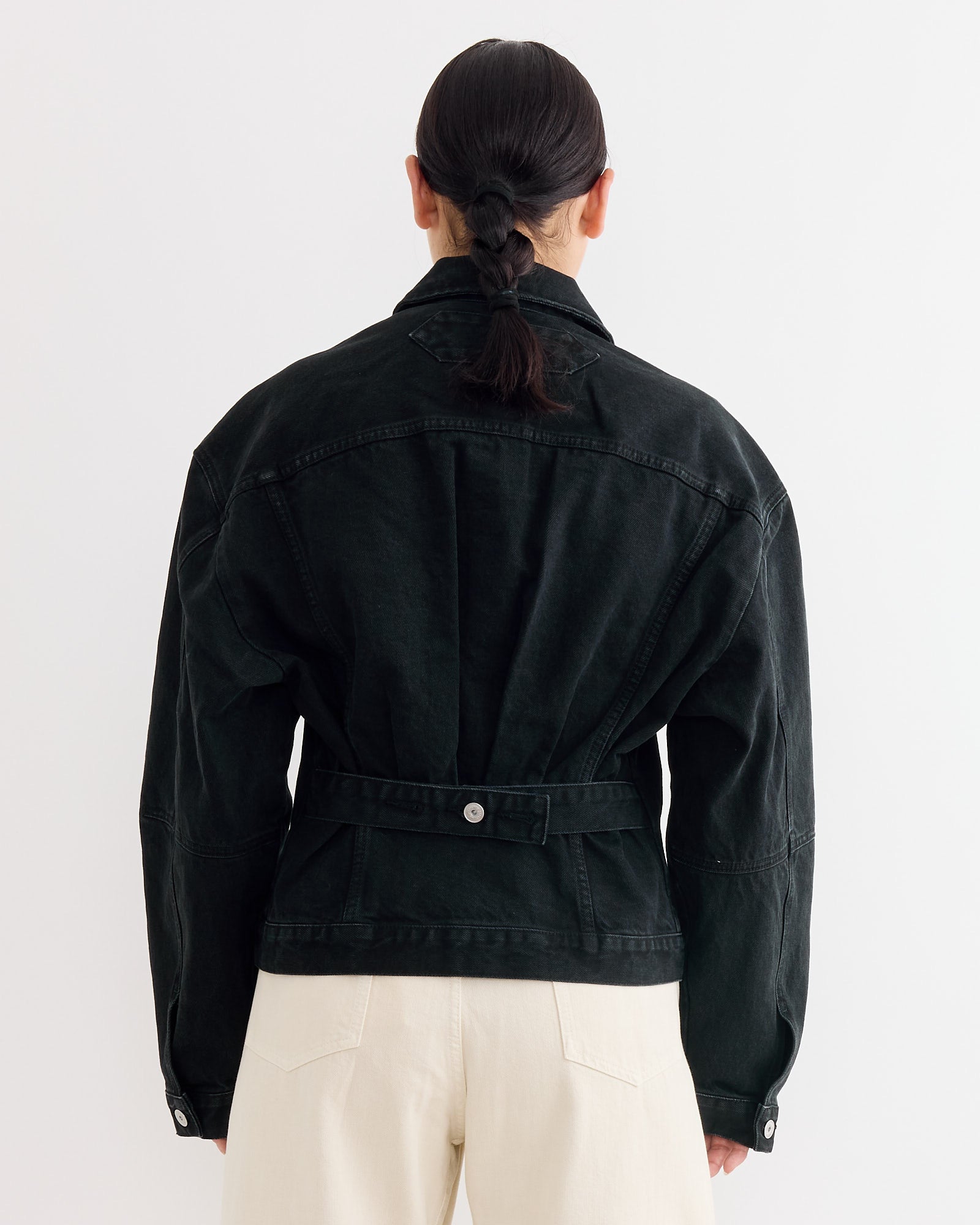A person with dark, neatly tied hair faces away, wearing the Jacquemus La Veste De-Nimes Jacket in Black and cream-colored pants—perfect inspiration for styling or checking your fit and sizing before shopping.
