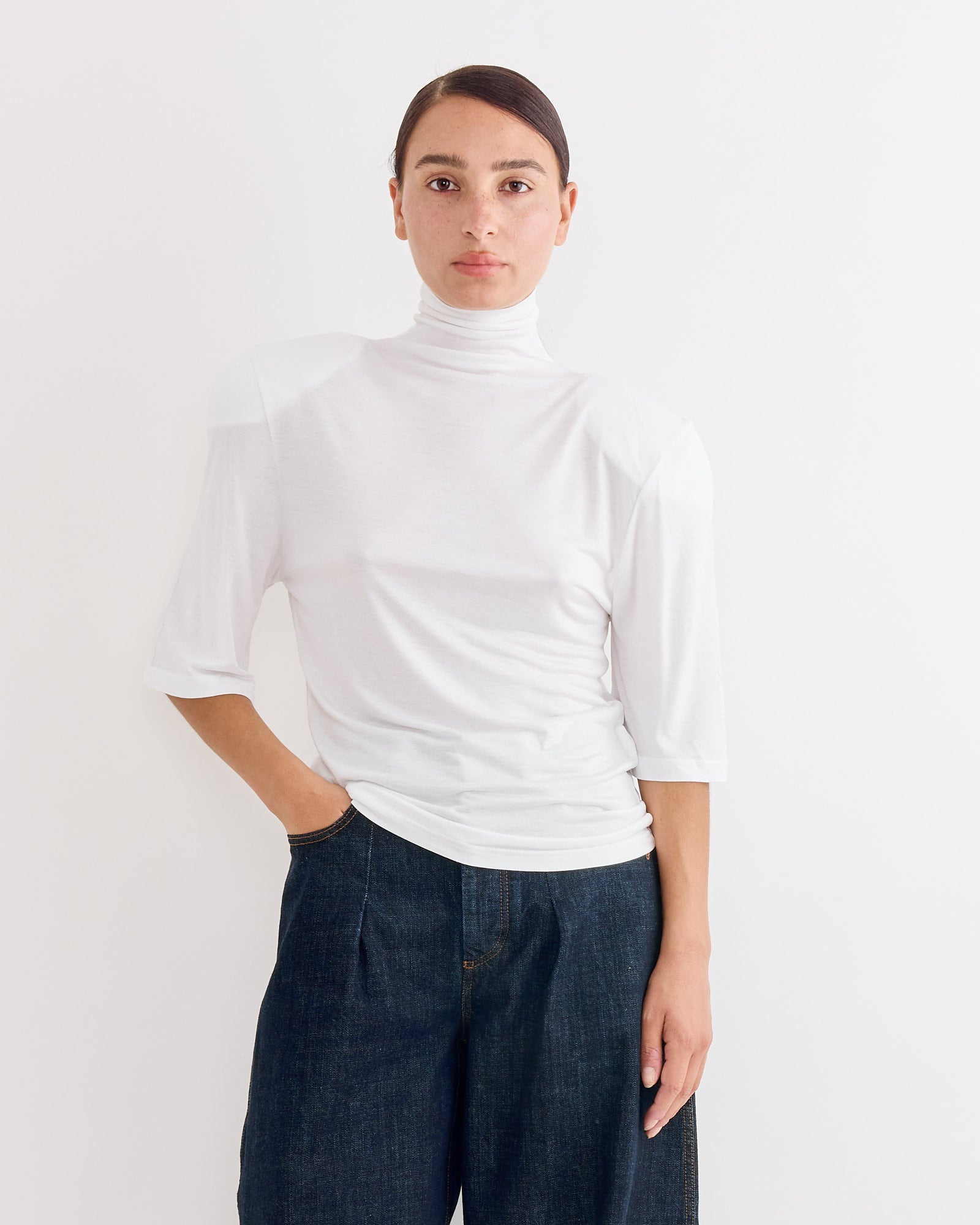 A person stands against a white backdrop, wearing the Jacquemus Le Spallina T-Shirt in White with elbow-length sleeves and dark wide-leg jeans. For fit or sizing questions, our online chat is here to help.