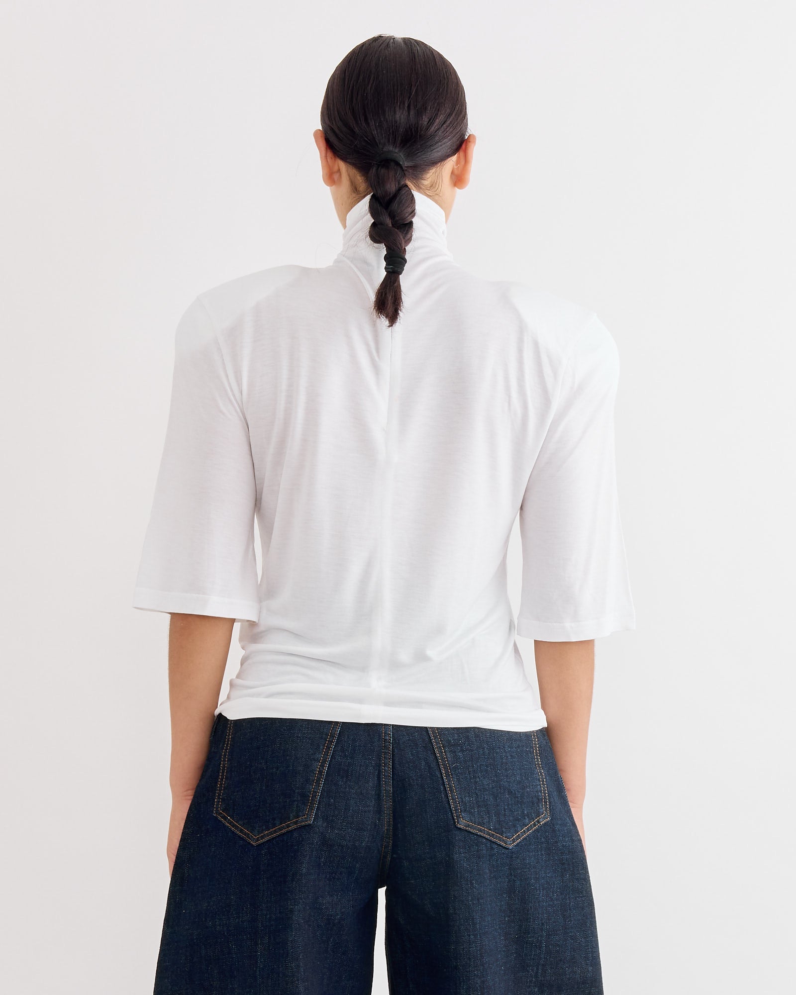 A person with dark hair in a braided ponytail is seen from the back, wearing the Jacquemus Le Spallina T-Shirt in White and dark high-waisted jeans against a plain white background, clearly displaying the fit and sizing.