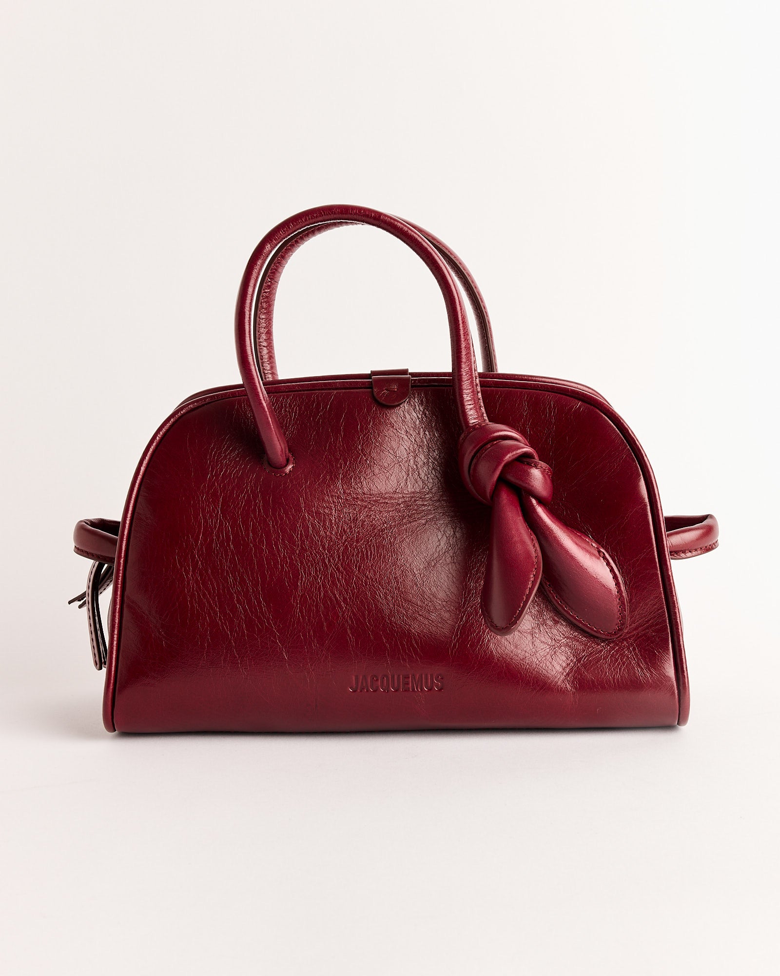 The Jacquemus Le Petit Turismo Bag features burgundy vintage-finished leather, two handles—one knotted—and a subtle embossed logo on the front, shown against a white background.
