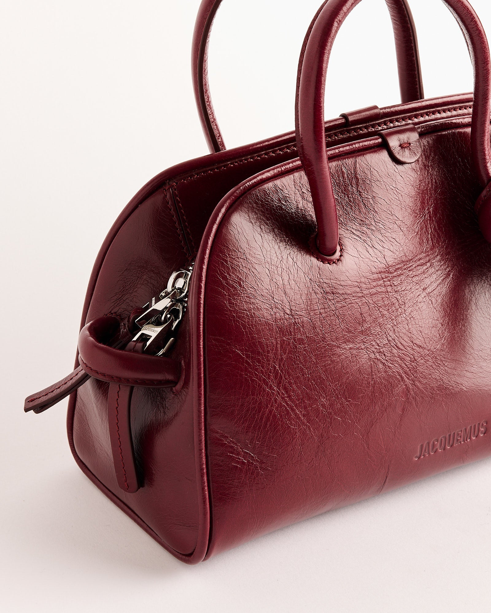 A close-up of the Jacquemus Le Petit Turismo Bag, a compact bowling bag crafted from structured burgundy vintage-finished leather with dual handles, a metallic zipper, and subtle embossed branding, set against a plain white background.