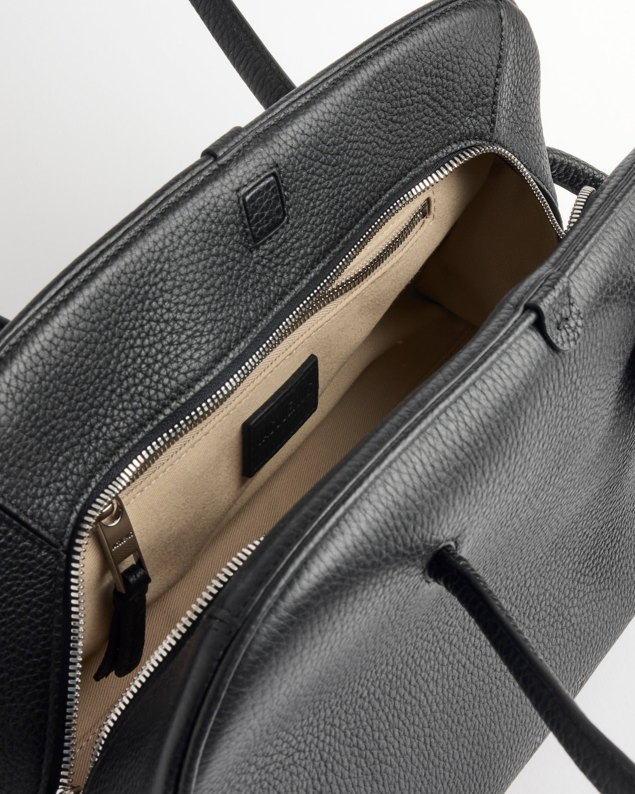 A close-up of an open Jacquemus Le Turismo Bag, showcasing its textured black leather exterior, beige interior, zippered pocket, small black label inside, and visible handles.