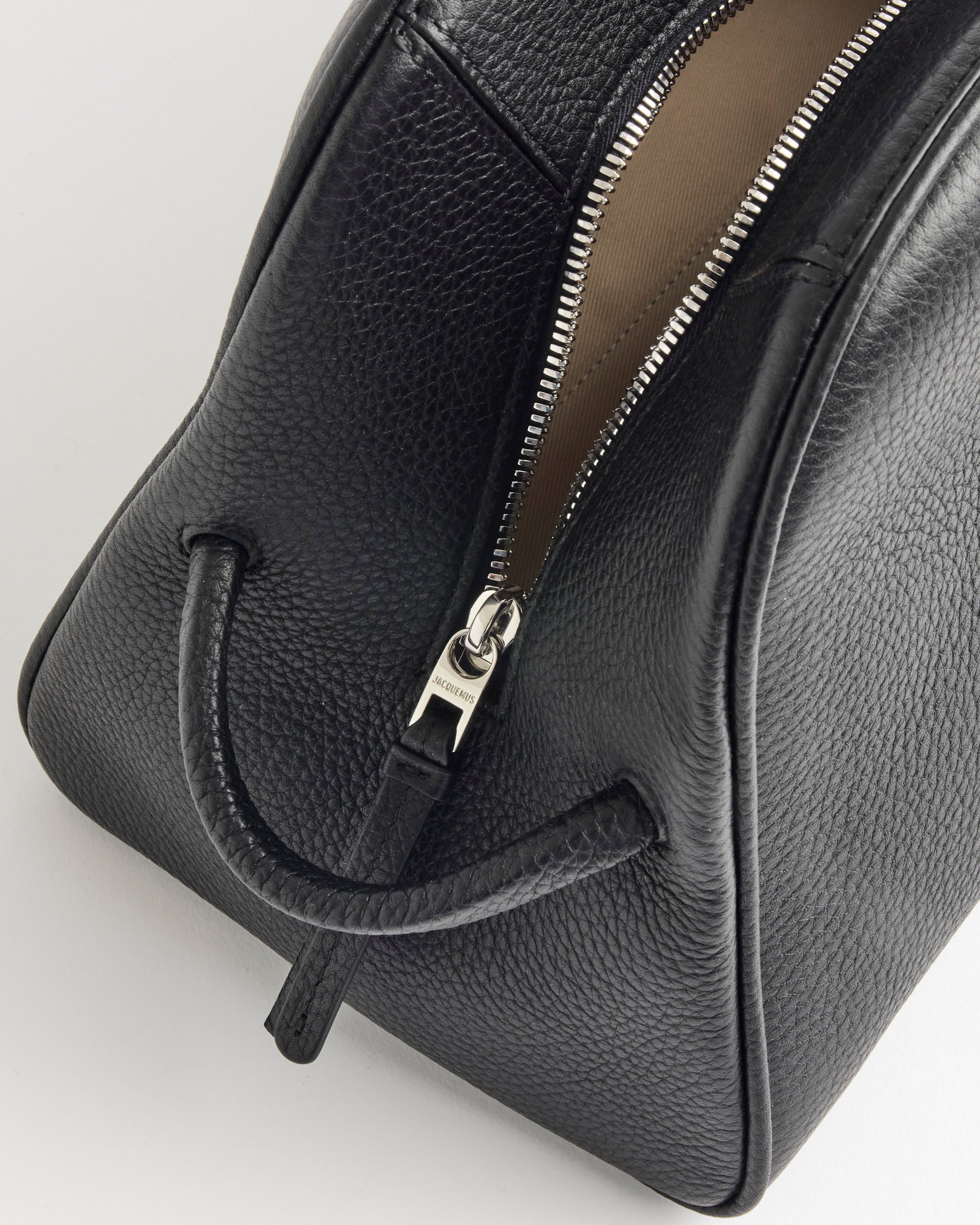 Close-up of the Jacquemus Le Turismo Bag, a black leather bowling bag with a pebbled texture, single curved handle across the front, and partially open zipper showing its beige interior.