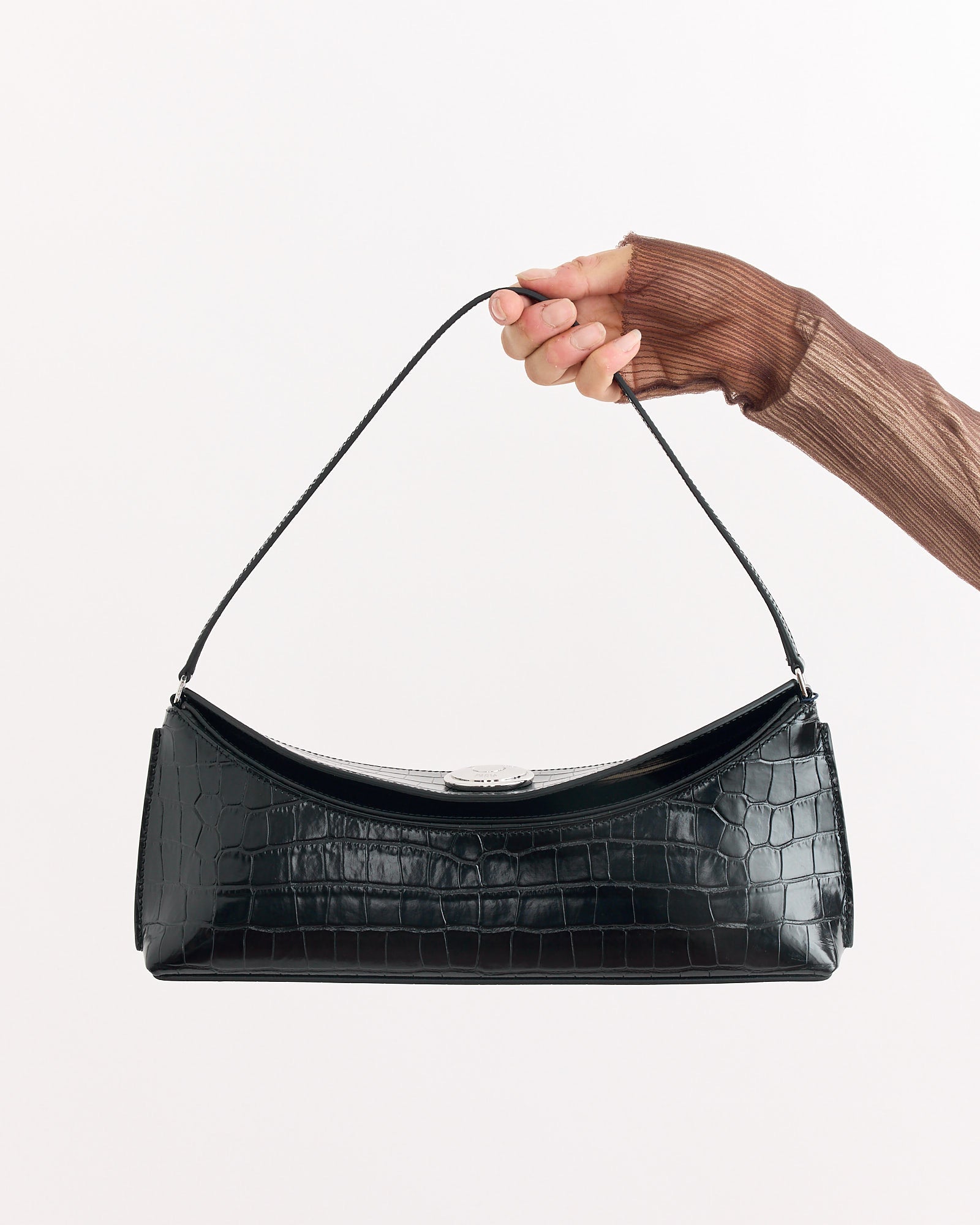 A hand in a sheer brown long-sleeve top holds the Jacquemus La Pochette Ovalo Bag in Black—a crocodile-embossed leather shoulder bag with a short strap—against a plain white background.