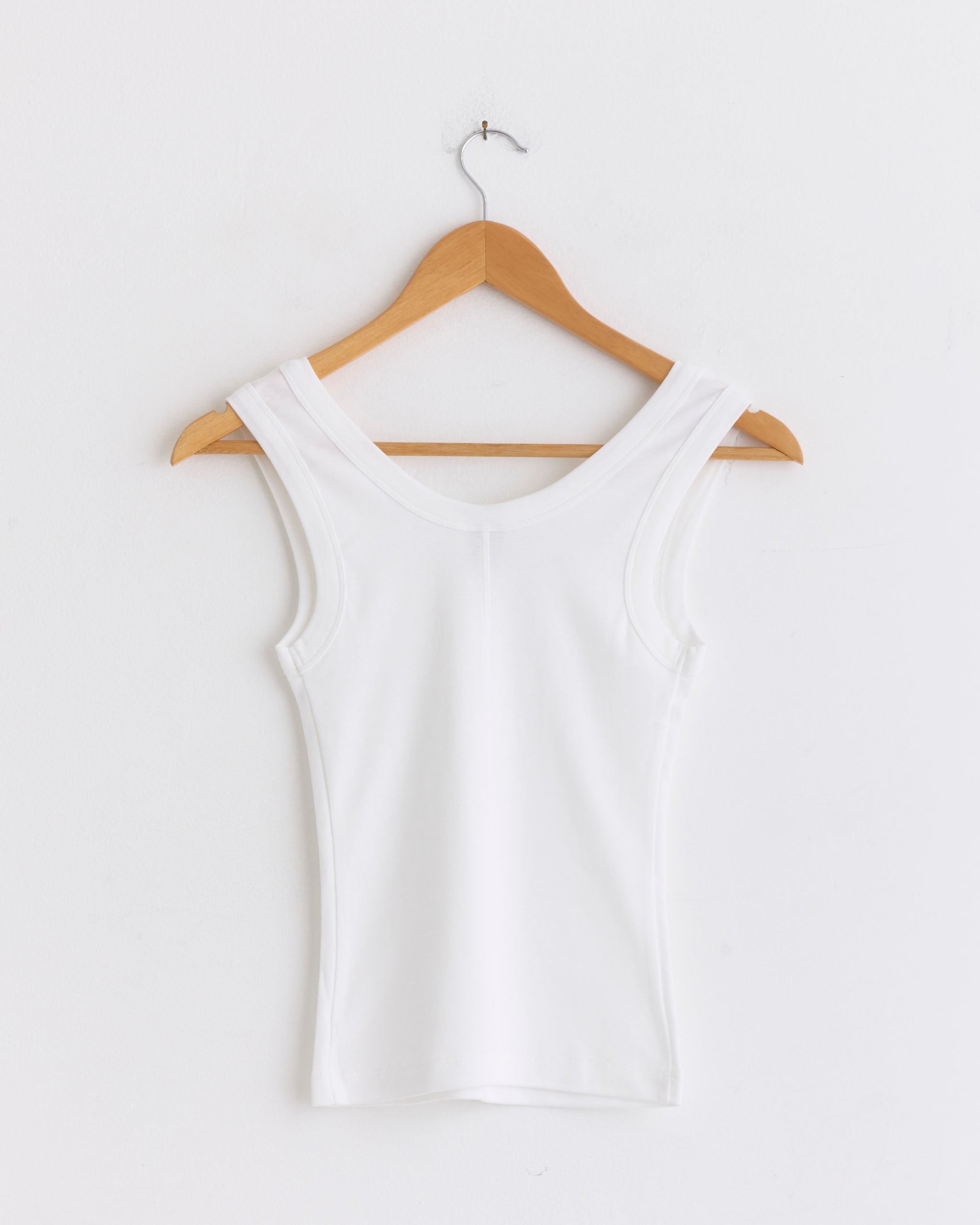 The Hillie Tank in White by FLORE FLORE, a women’s scoop neck top made from soft organic cotton, hangs on a wooden hanger against a plain white wall.
