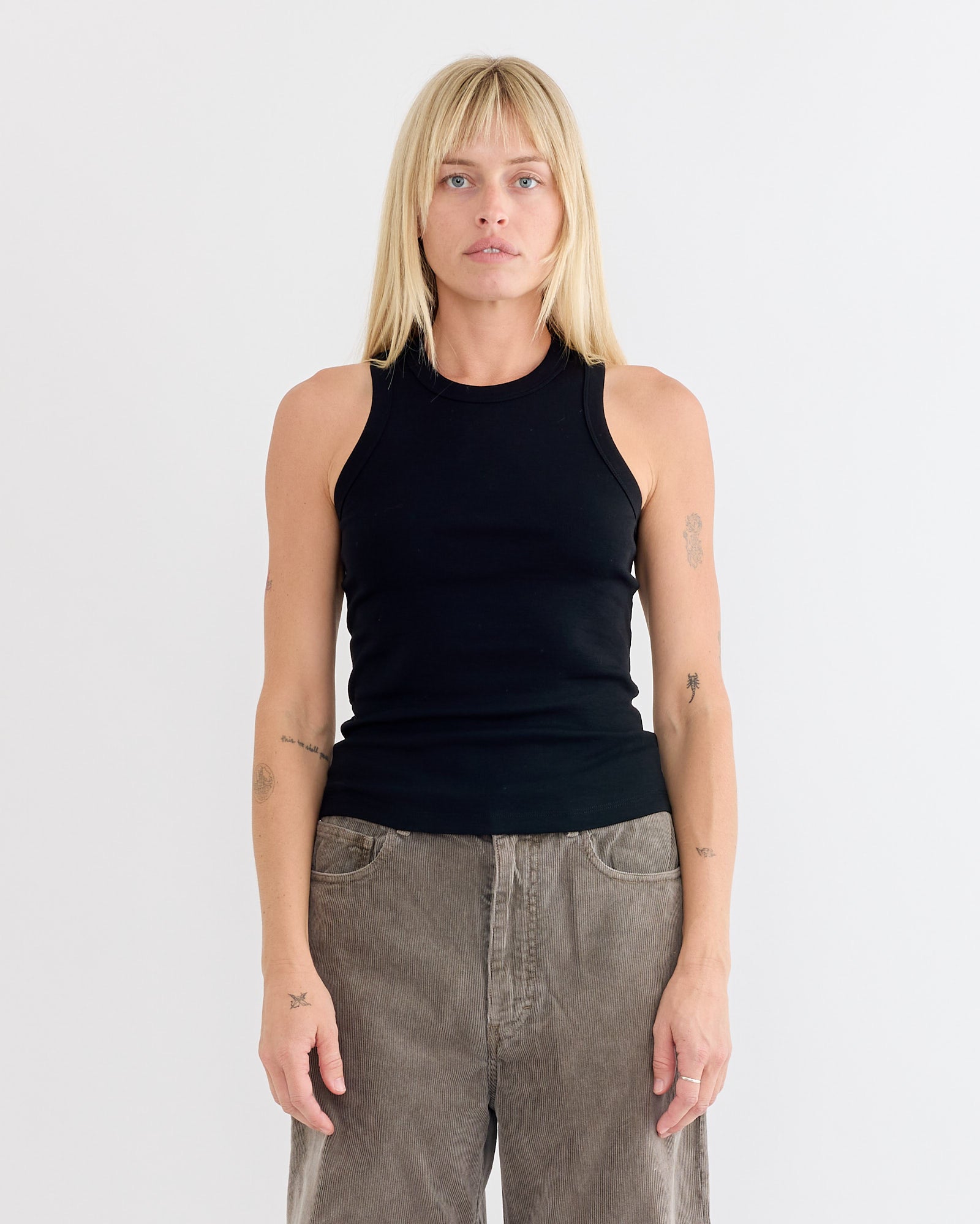 A person with straight blonde hair and bangs wears the FLORE FLORE Hannah Tank in Black with loose brown pants, standing against a plain white background with a neutral expression.