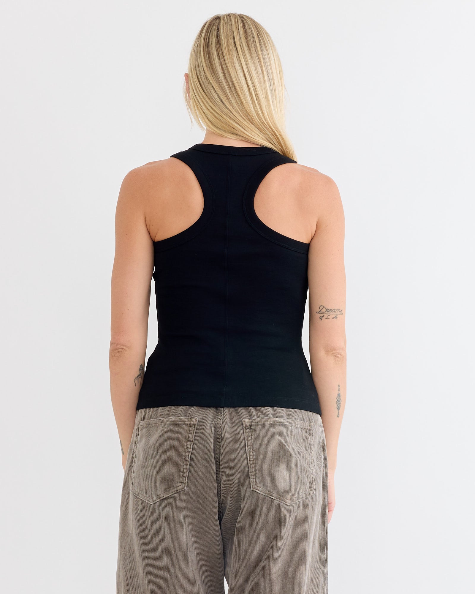 A person with long blonde hair is seen from the back, wearing the FLORE FLORE Hannah Tank in Black and loose gray pants, standing against a plain white background.