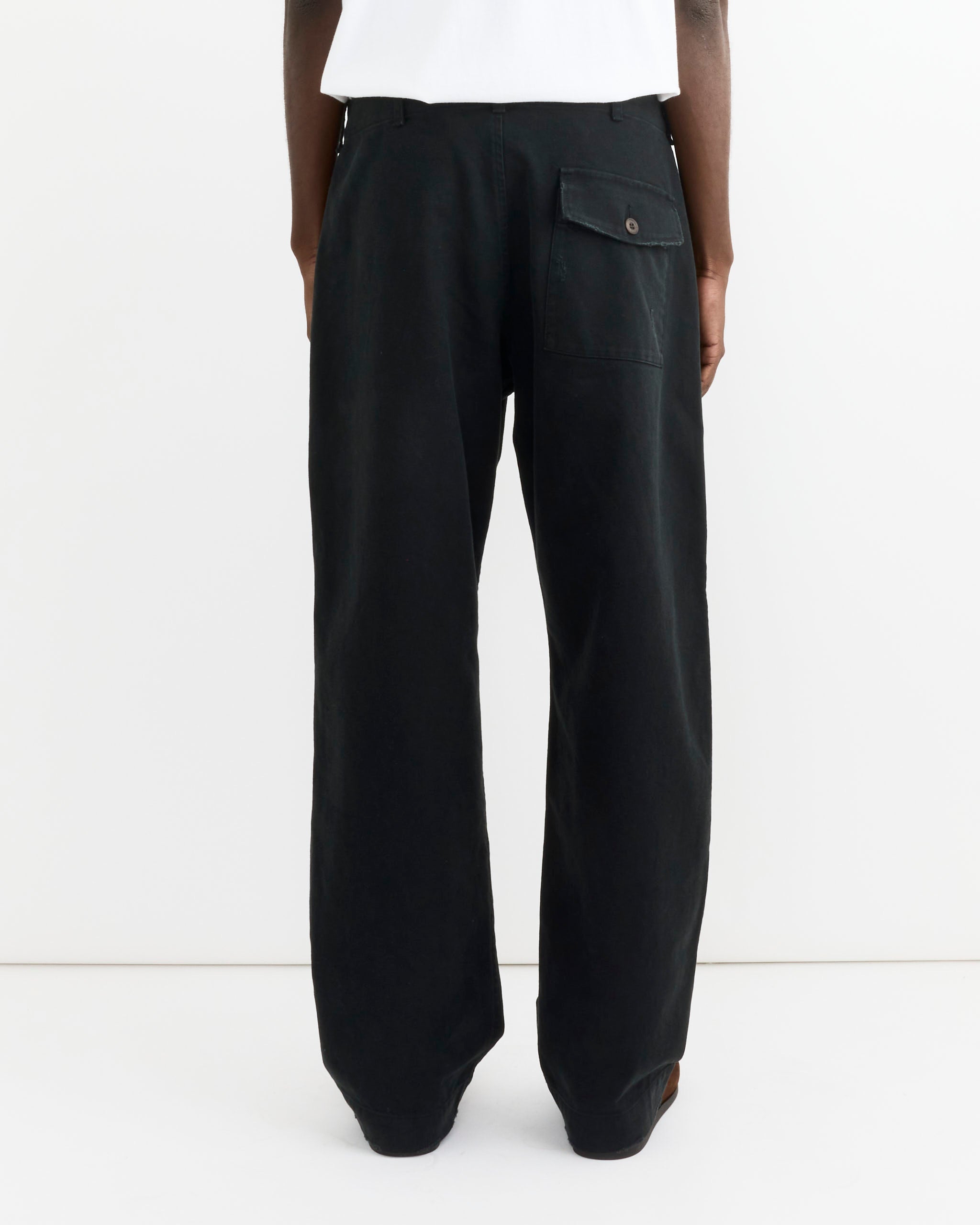 A person stands against a white background, seen from behind wearing SMOCK's Toyon Pant in French Cotton Twill Black, featuring relaxed straight legs and a button-flap back pocket, paired with a white top. Only the lower half is visible.