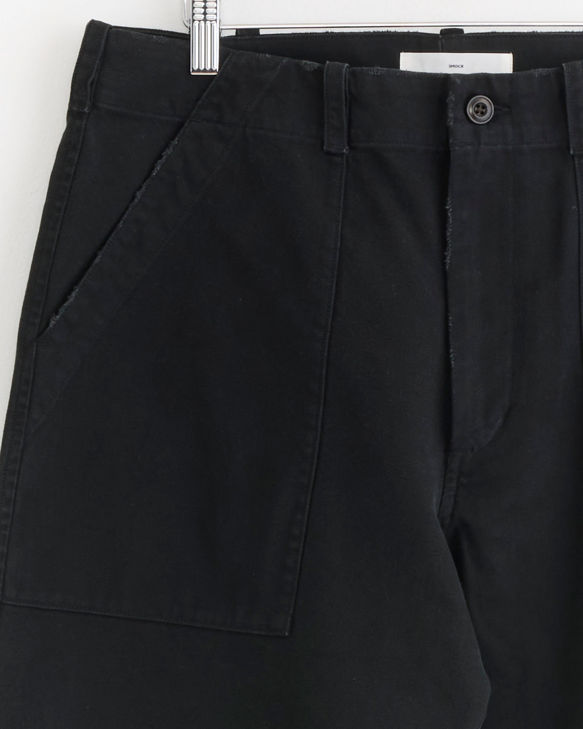 Close-up of SMOCK Toyon Pant in French Cotton Twill Black, with large front pockets, button closure, visible belt loops, and a relaxed straight-leg fit, hanging on a hanger against a white background.