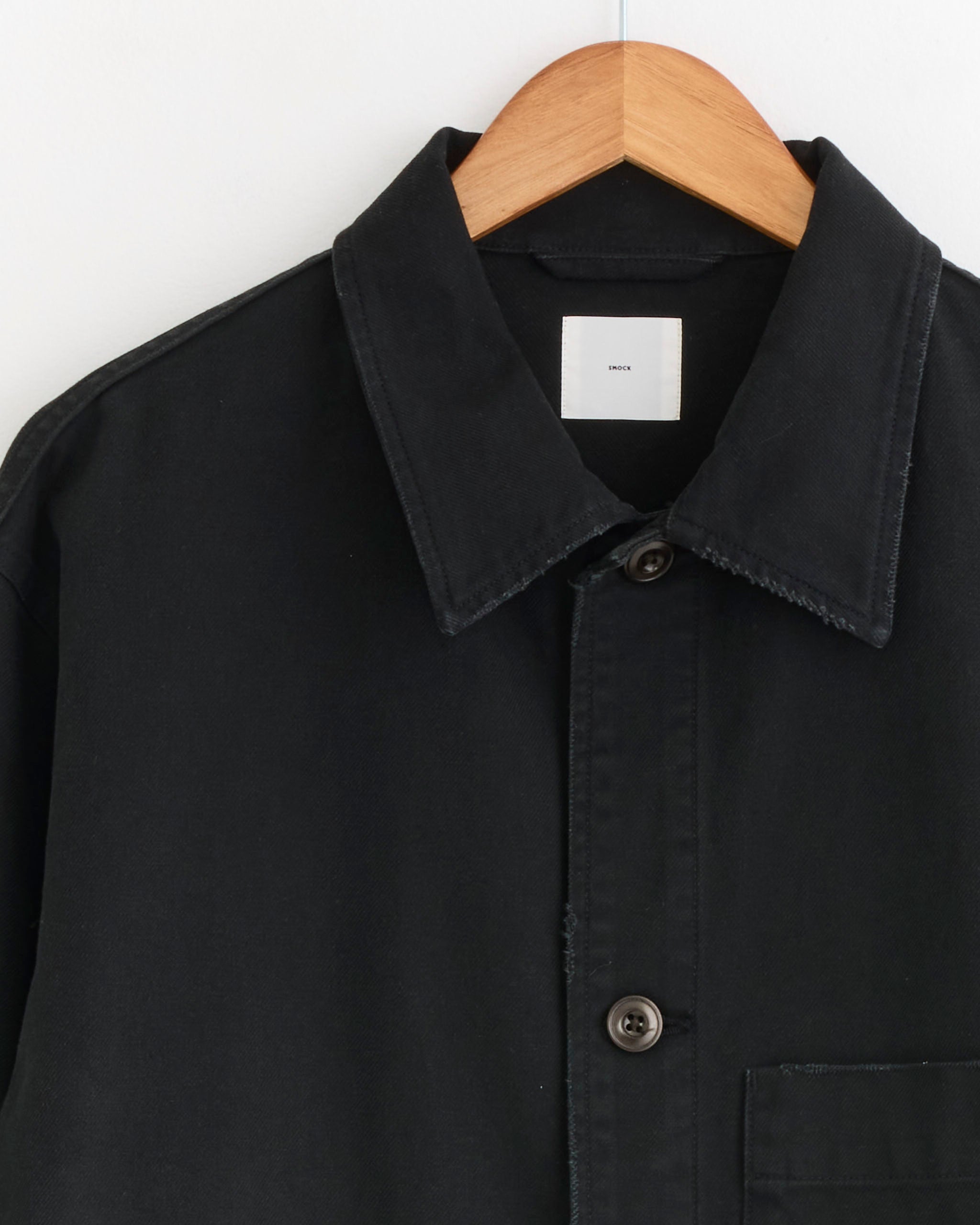 The SMOCK Lang Jacket in French Cotton Twill Black hangs on a wooden hanger against a white background, showing only the upper portion with two visible buttons and part of a chest pocket.
