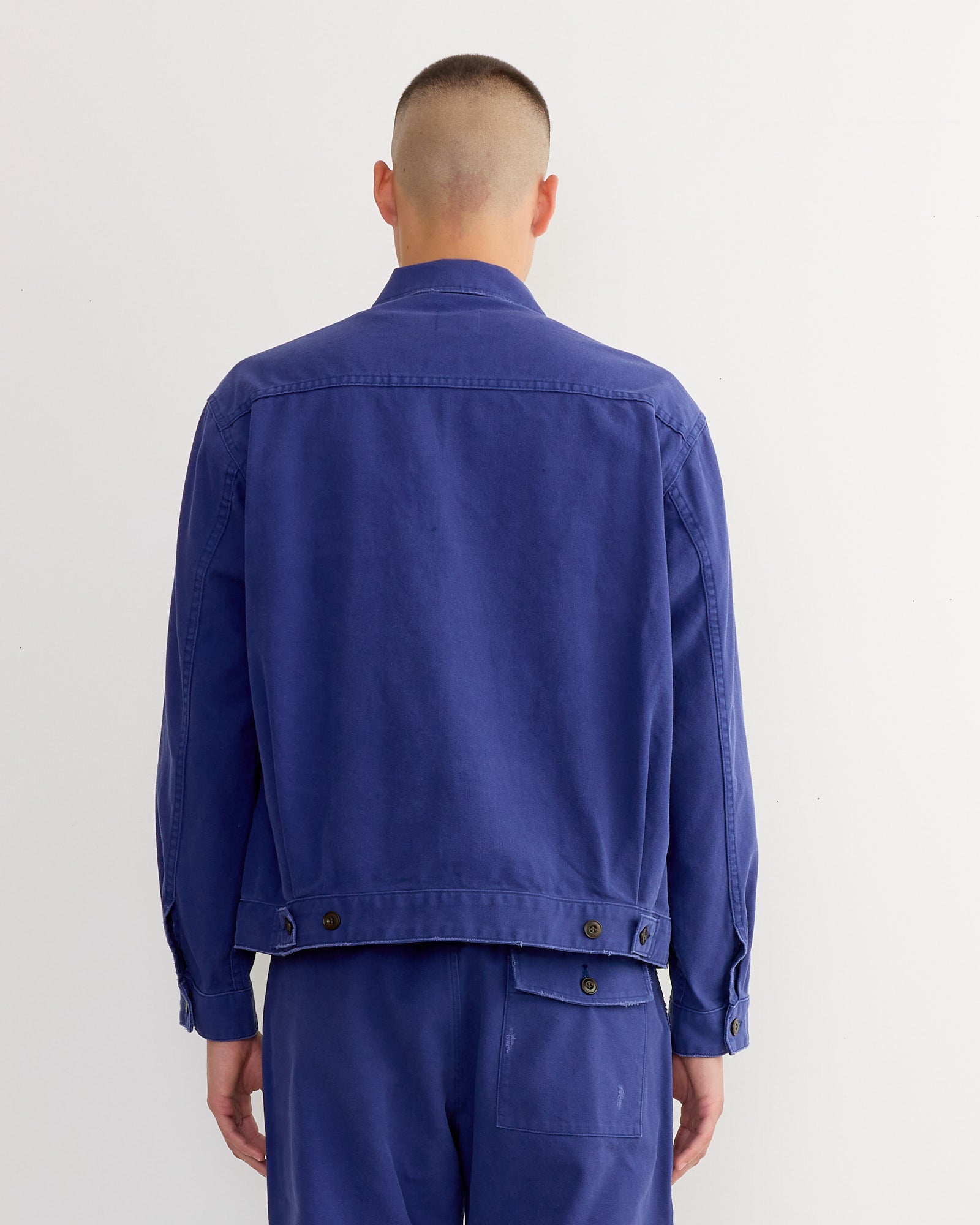 A person with a buzz cut faces away from the camera, wearing the SMOCK Lang Jacket in French Cotton Twill Blue and matching denim pants, both crafted from Japanese-woven French cotton twill, set against a plain white background.